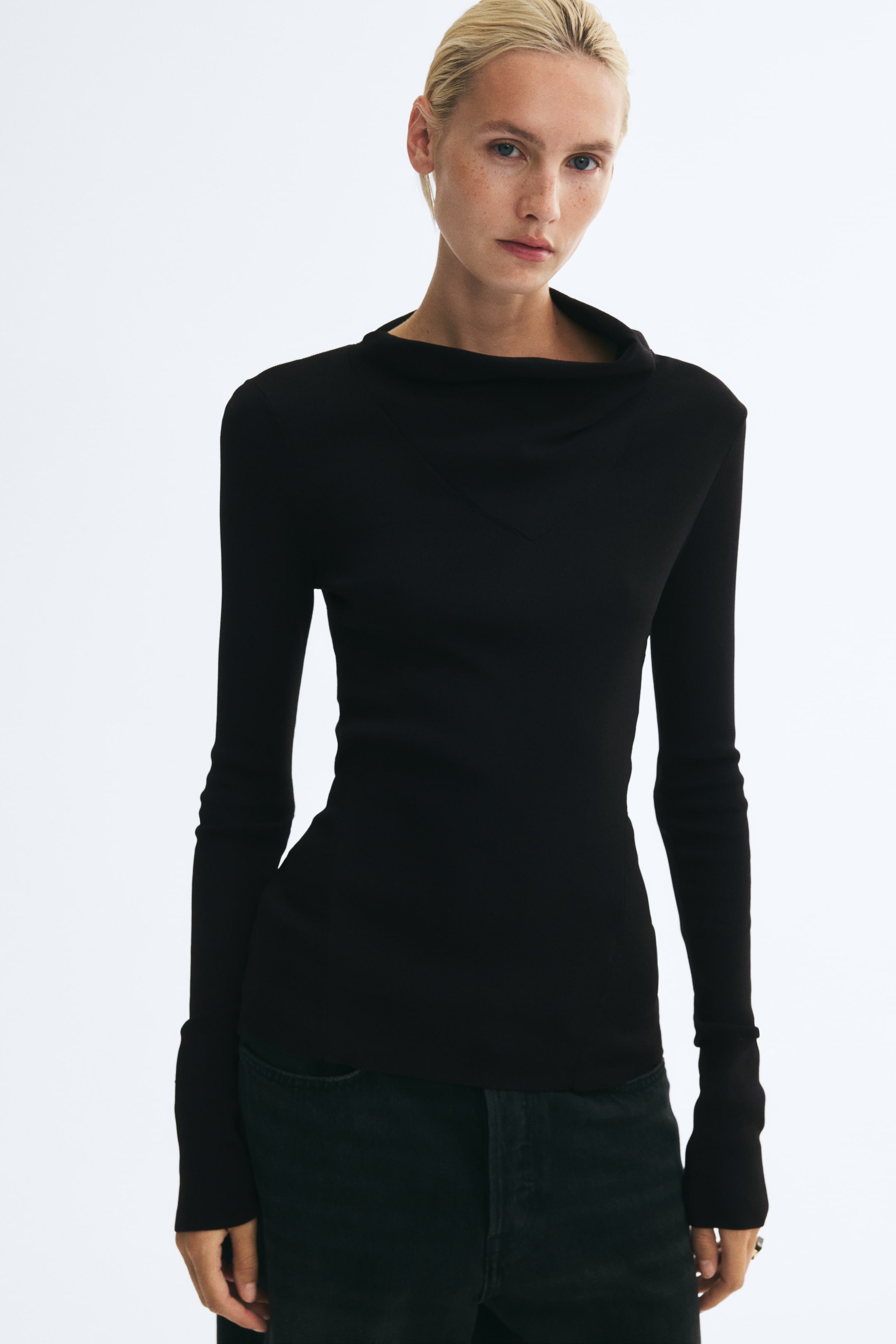 View larger image: A black, fitted rib-knit top featuring an asymmetrical fold-over collar and a V-shaped seam at the chest is worn by the model. The long-sleeved top has shaping seams down the front and is styled with dark-wash jeans.