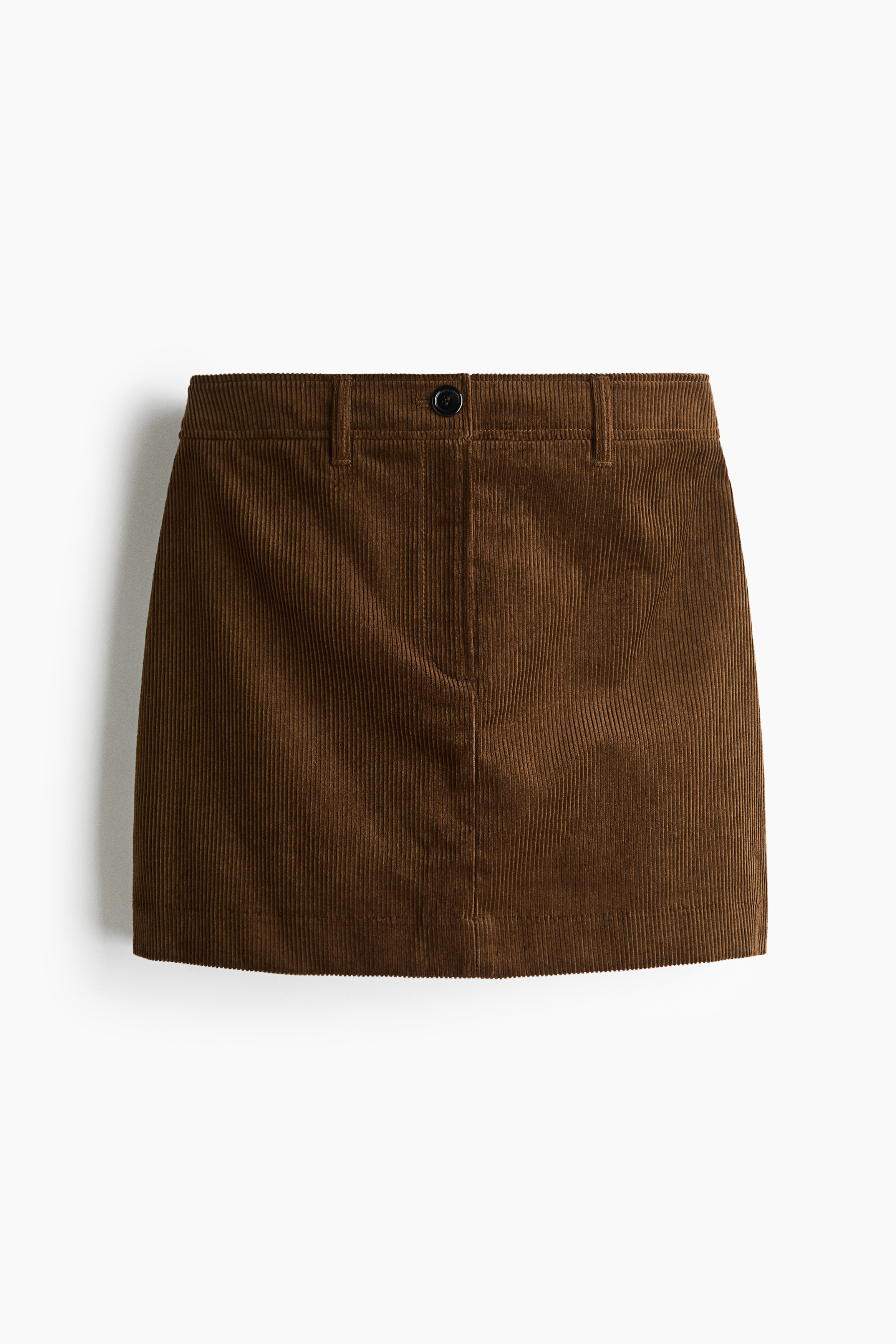 The model is wearing a brown corduroy mini skirt, styled with a dark belt at the waist. The skirt is paired with a light blue button-down shirt and a grey knit sweater draped over the shoulders.