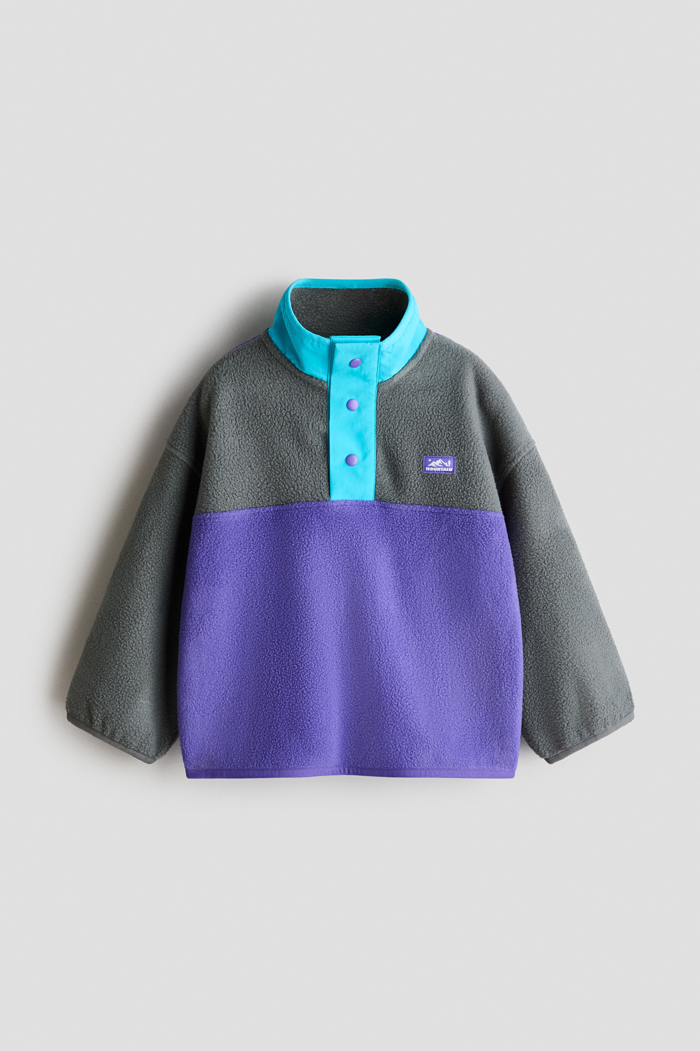 A colour-blocked fleece sweater featuring a grey upper section, a purple lower section, and a turquoise stand collar with a press-stud placket is worn by the model. The sweater is styled with straight-leg blue jeans and white trainers.