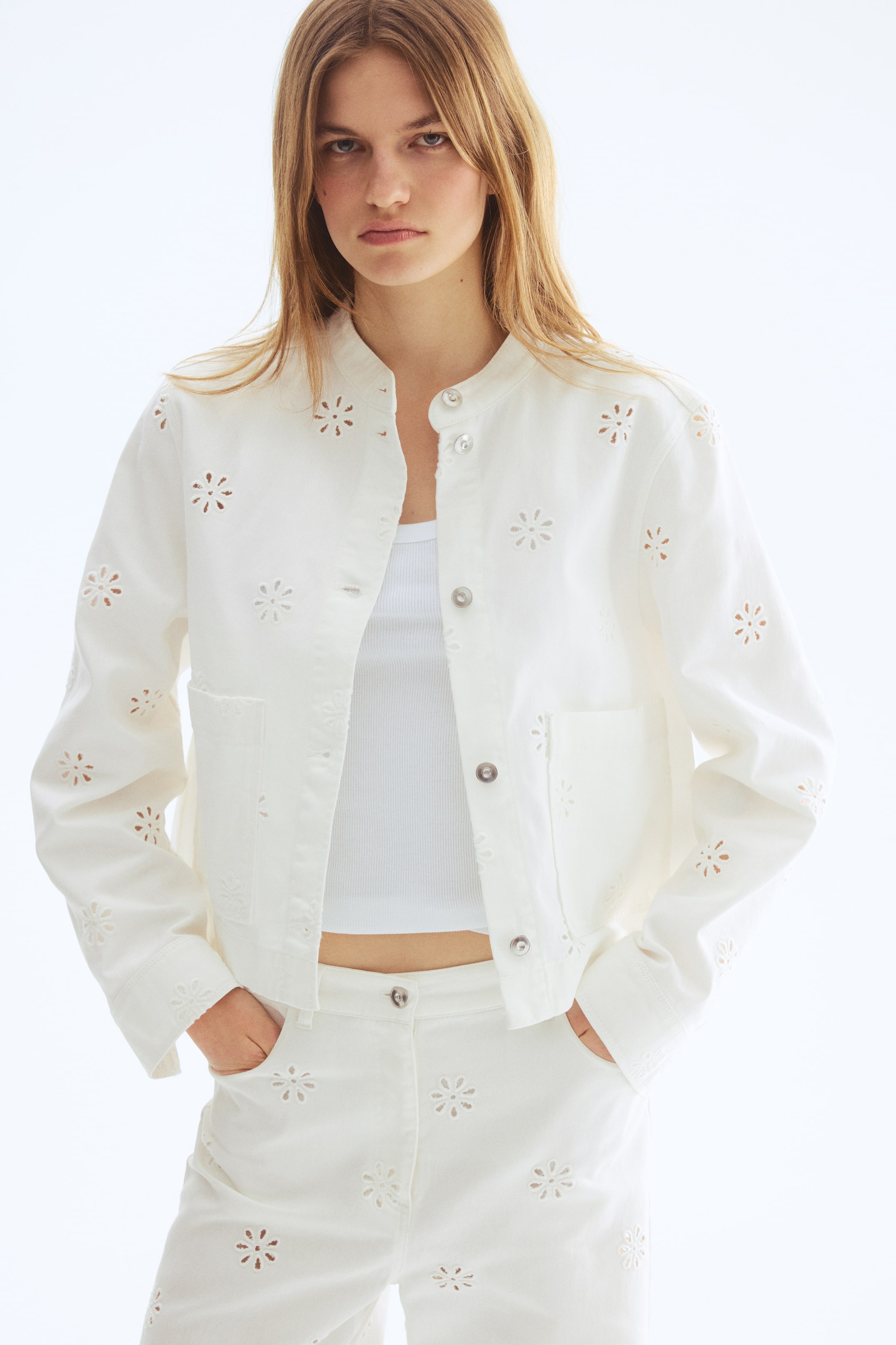 View larger image: Presenting a white eyelet jacket paired with coordinating trousers, displayed facing forward. The jacket boasts a band collar, silver-toned snap buttons, and two patch pockets, all embellished with intricate cut-out floral patterns. These eyelet designs beautifully extend to the matching trousers, creating a cohesive look.