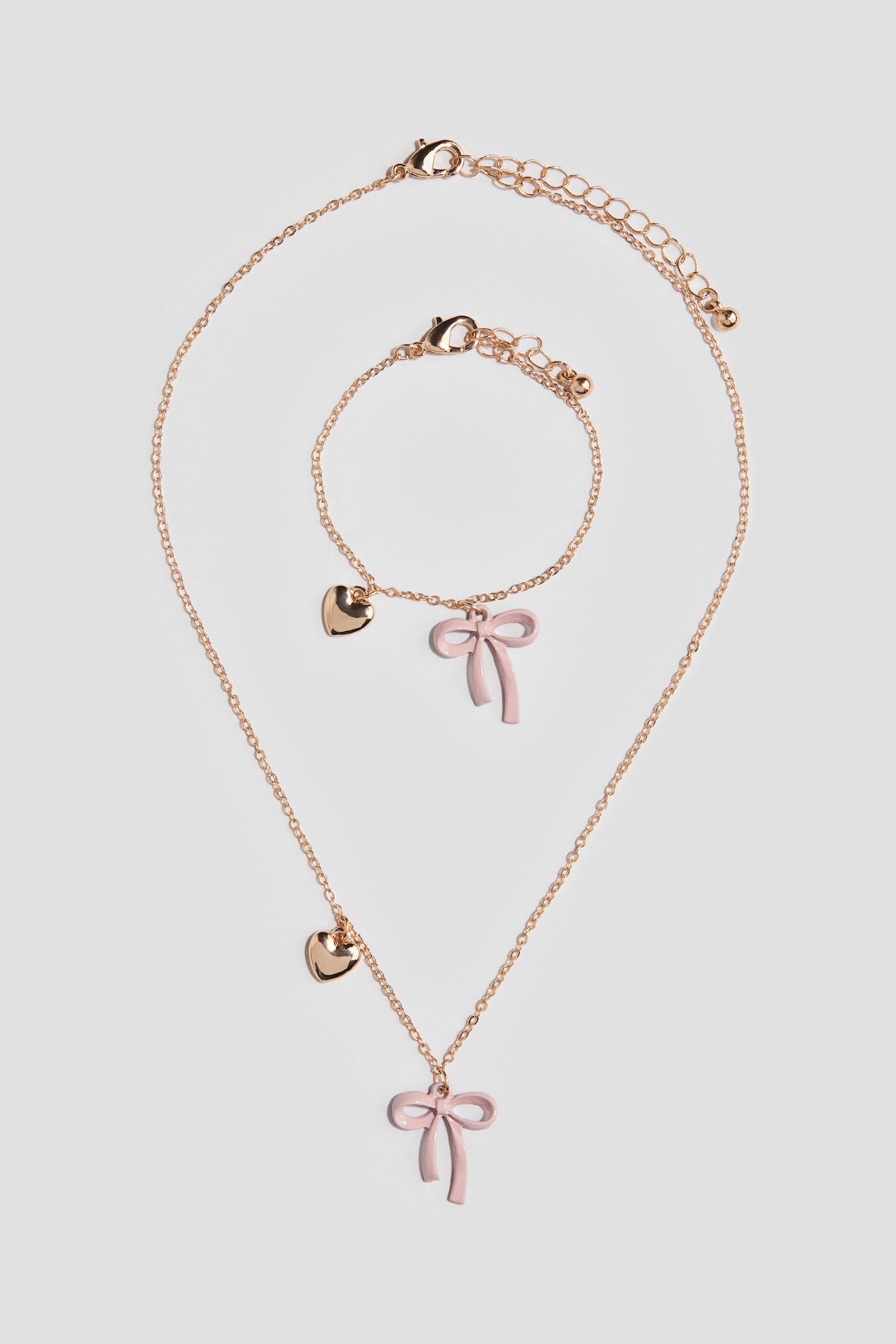 Un set di gioielli color oro mette in mostra una collana coordinata e un bracciale. Entrambi i pezzi presentano una catenina a maglia fine con chiusura regolabile a moschettone, un piccolo charm a cuore lucido color oro e un pendente a fiocco smaltato di rosa chiaro. - 1