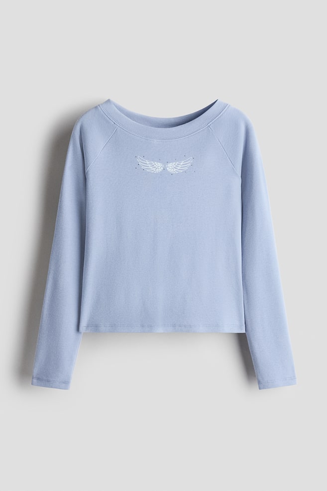 A light blue, long-sleeved top is shown from the front, featuring a crew neck, raglan sleeves, and a straight hem. A small white graphic of angel wings with tiny surrounding dots decorates the centre of the chest. - 1