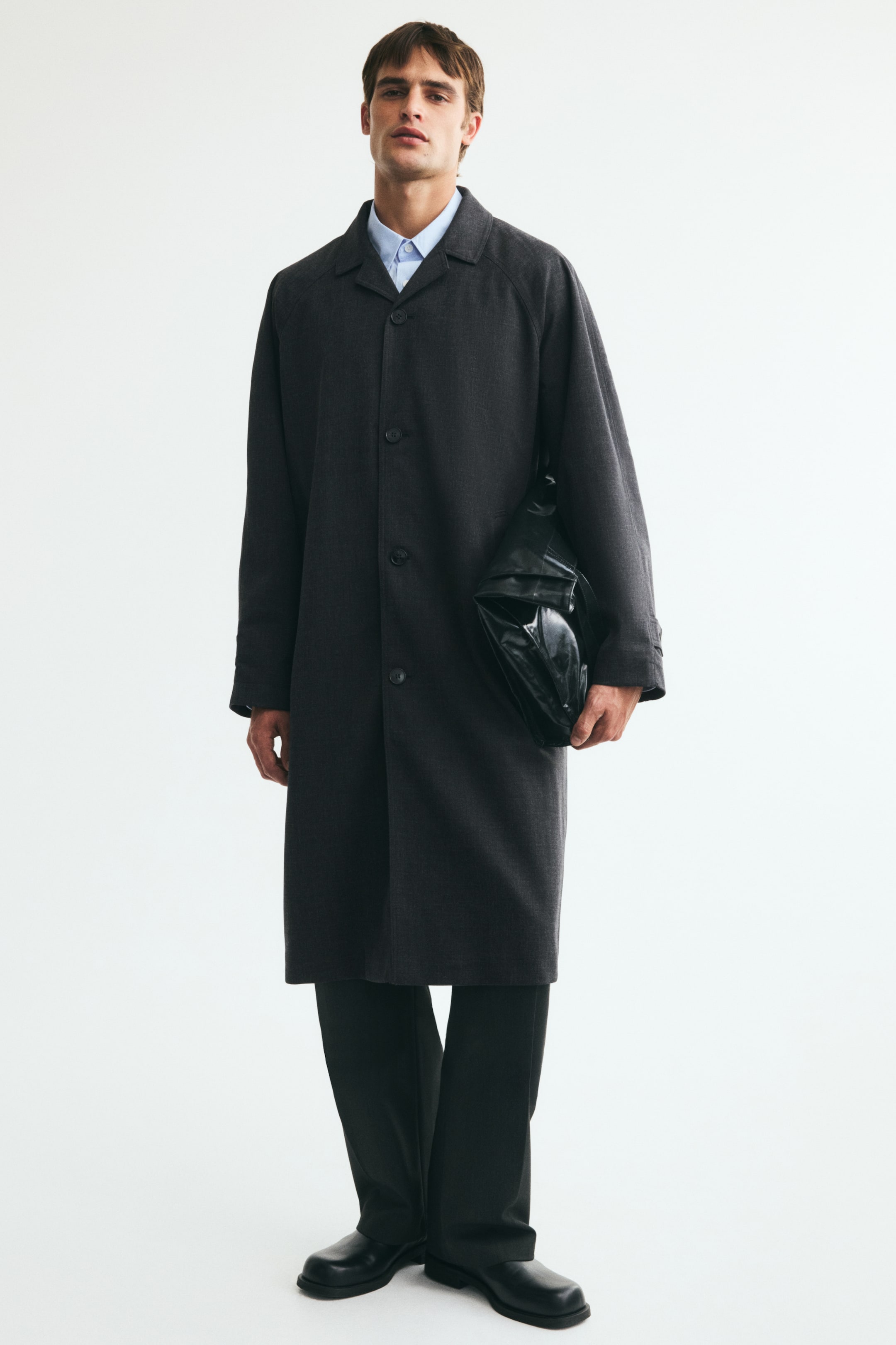 View larger image: A dark grey car coat in twill is being worn by the model, featuring a classic collar, a button-down front, and long raglan sleeves. The coat is styled over a light blue collared shirt and dark trousers, while the model is holding a black bag under one arm.