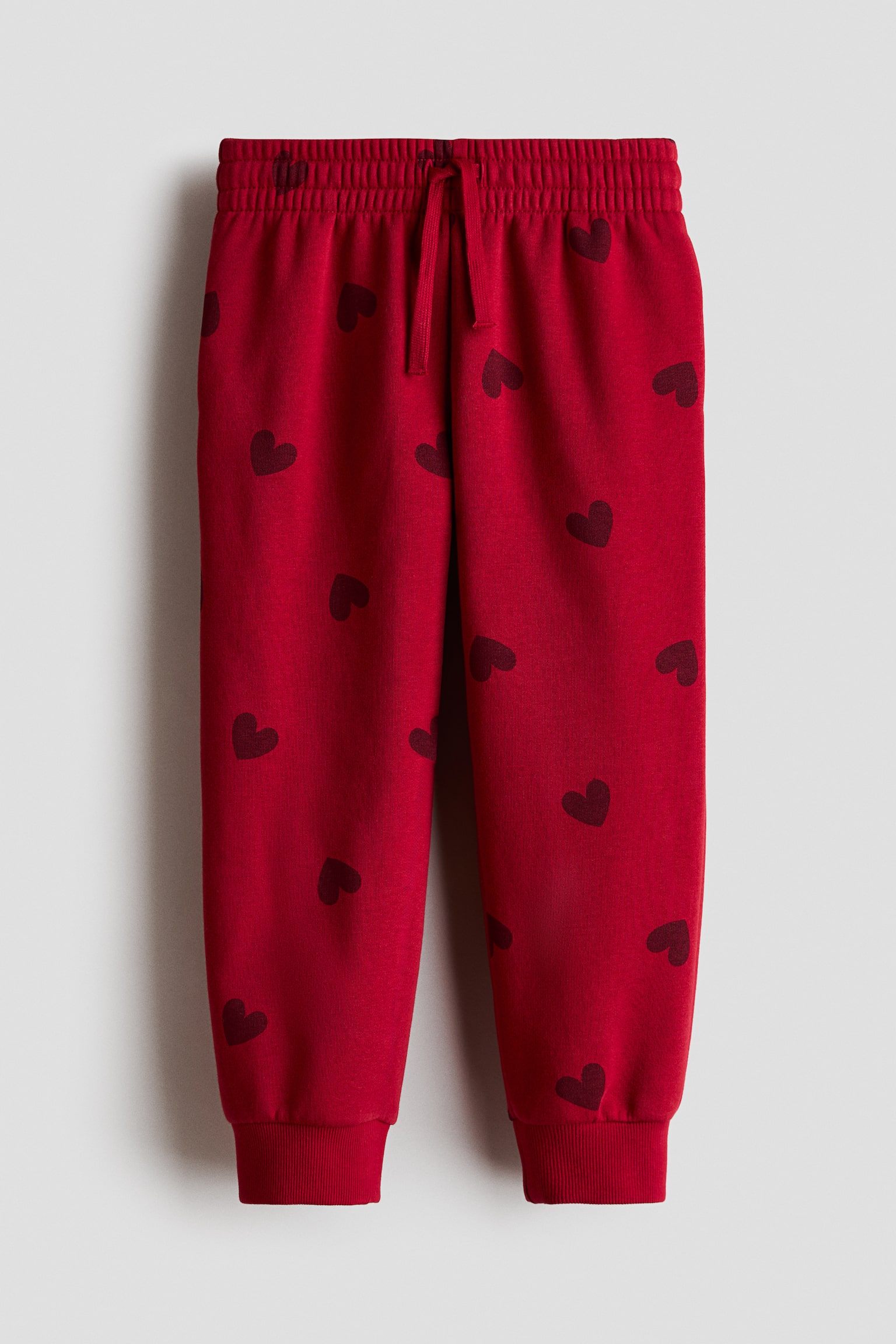 Pantaloni de trening roÈii, cu model integral de inimioare roÈu Ã®nchis, sunt prezentaÈi din faÈÄ, evidenÈiind o talie elasticatÄ cu Ènur asortat Èi manÈete striate.