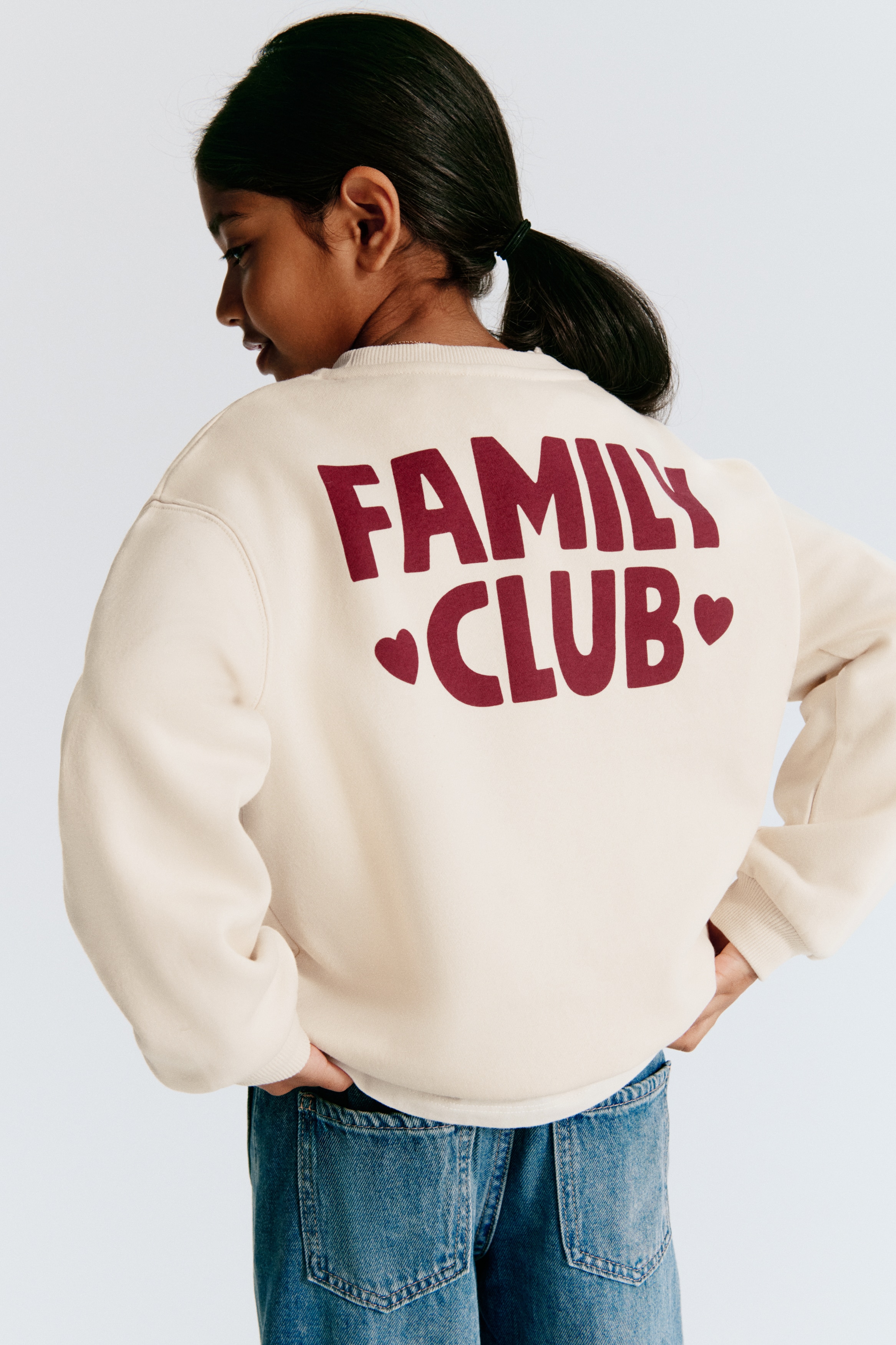 A cream-coloured sweatshirt is shown from the front, featuring a round, rib-trimmed neckline, dropped shoulders, and ribbed cuffs and hem. The chest area showcases a dark red graphic print that reads "FAMILY CLUB" with a small heart detail dotting the letter "I."