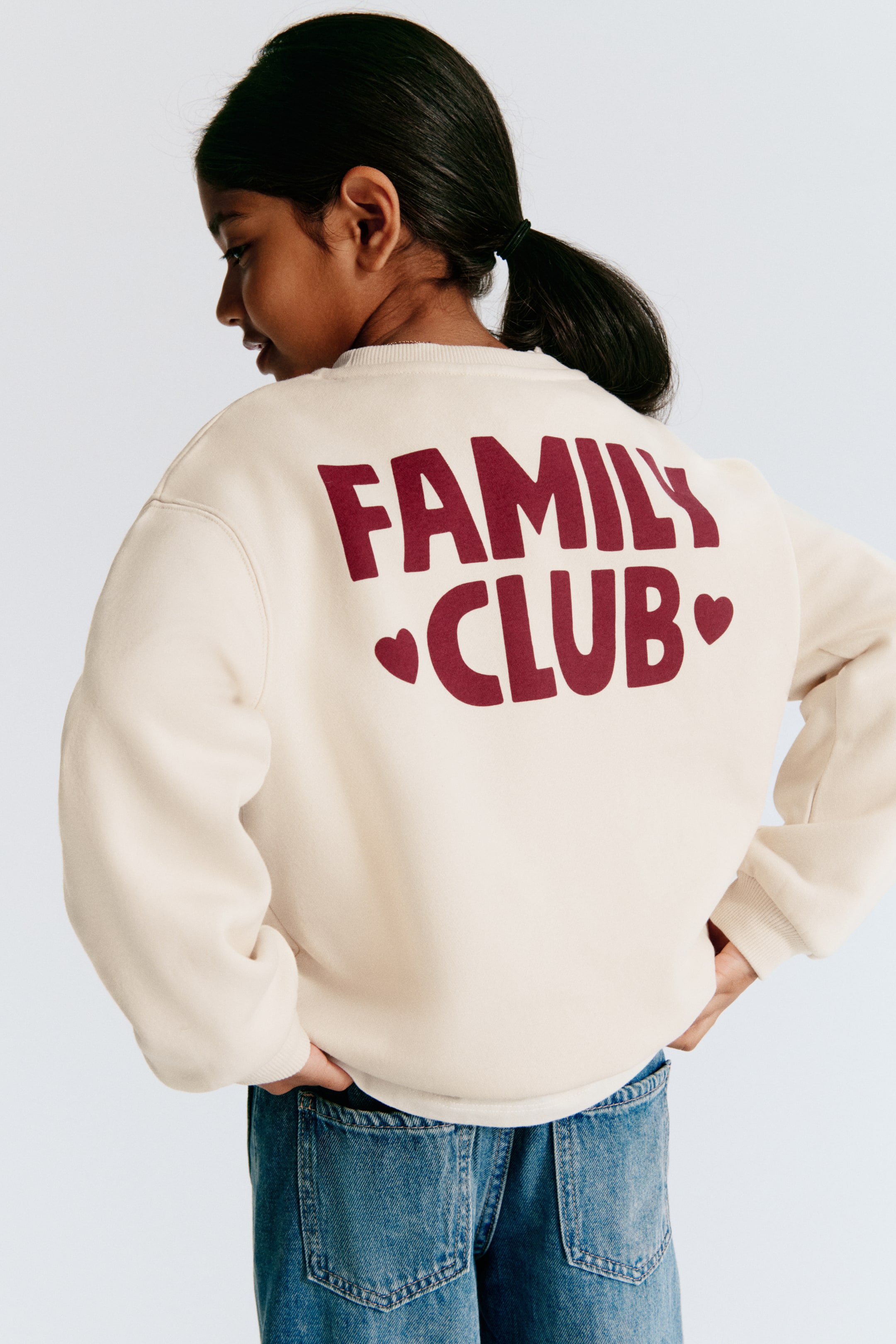 View larger image: An off-white sweatshirt is shown from the back, featuring a relaxed fit with dropped shoulders, ribbed neckline, ribbed cuffs, and a ribbed hem. The back of the sweatshirt features a large burgundy graphic print reading "FAMILY CLUB", flanked by two small hearts.
