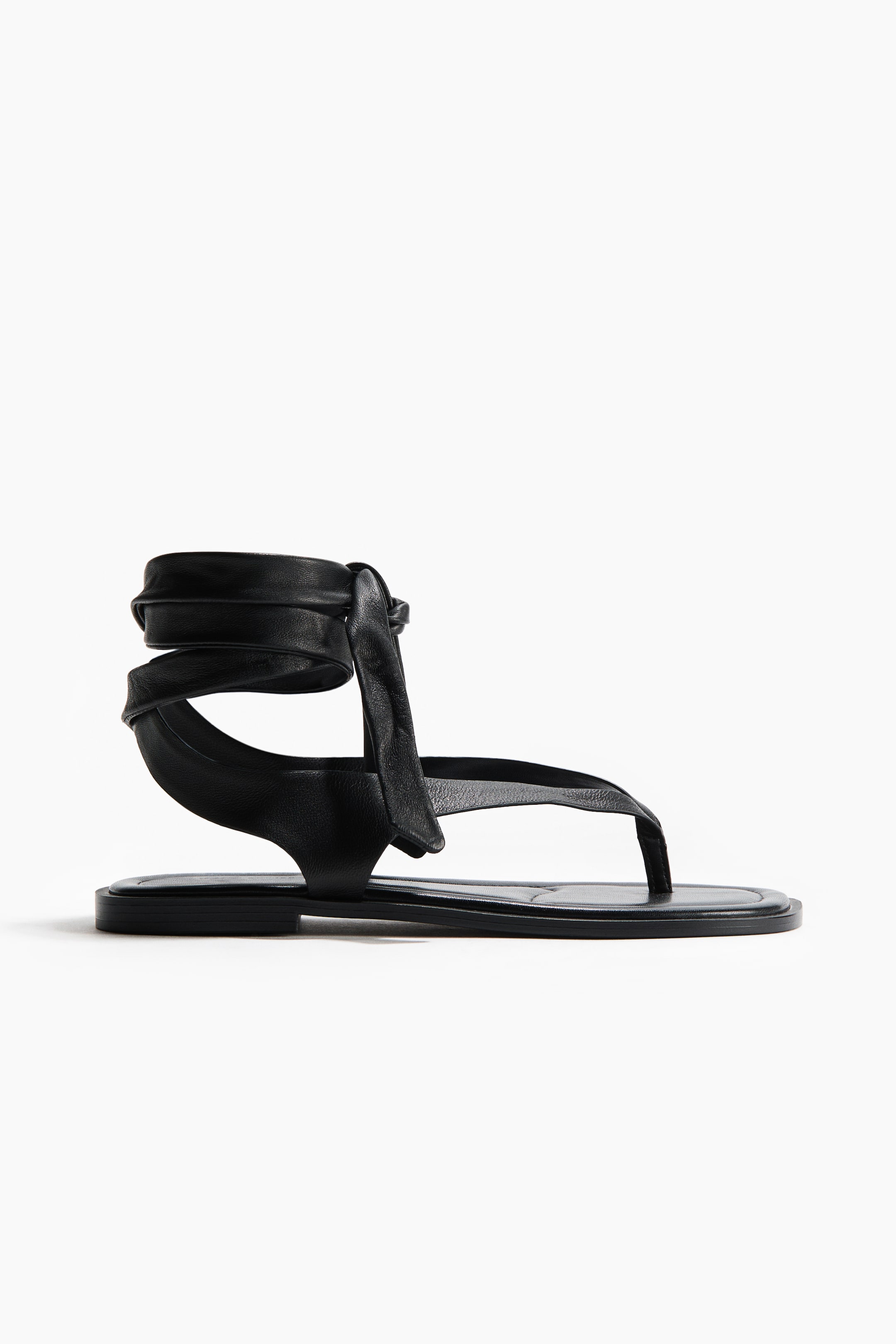 View larger image: A black flat sandal featuring a thong toe strap that connects to a wide side strap, curving upwards to form a soft, gathered ankle wrap, tying into a knot on the side.