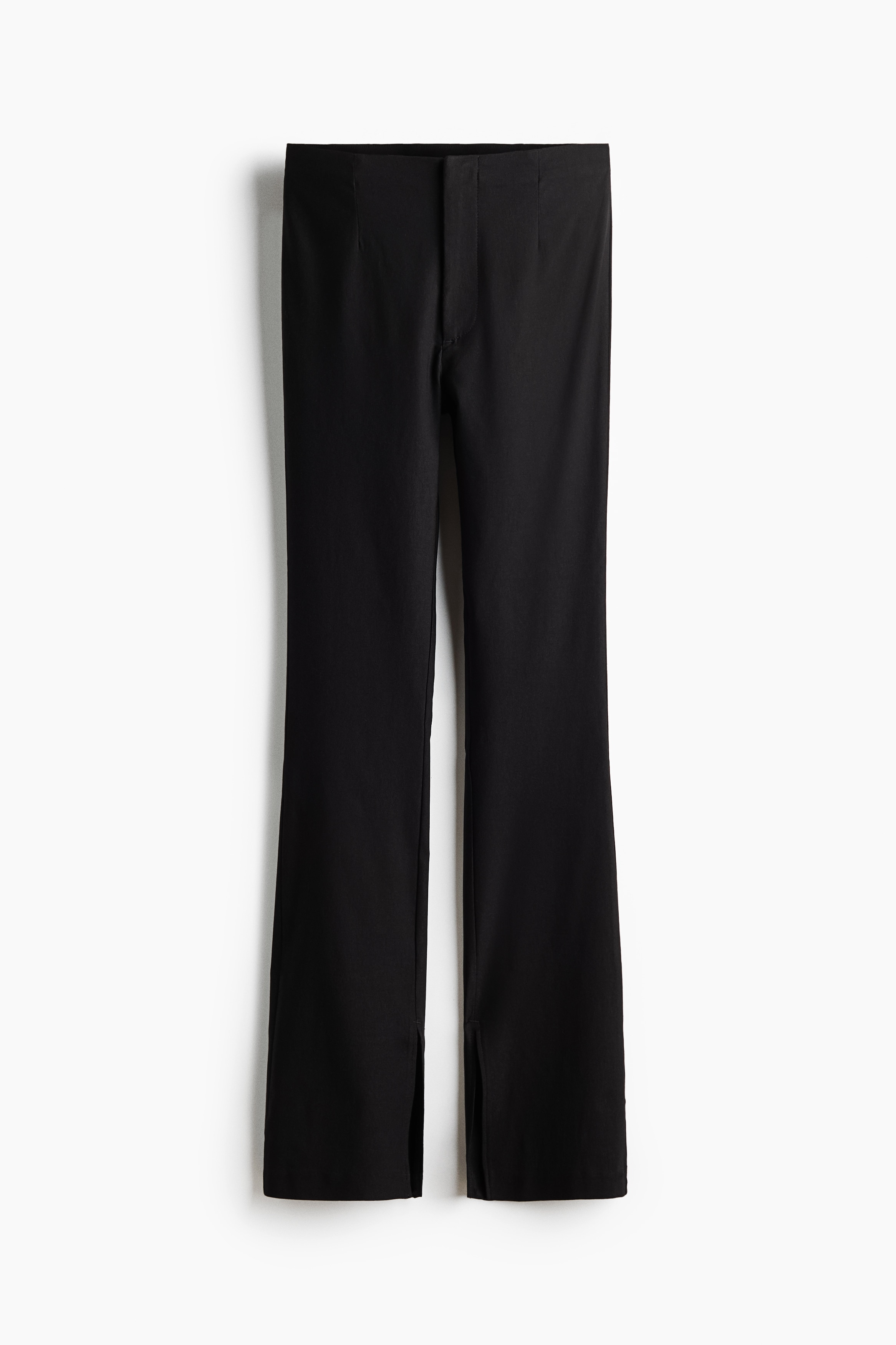 Black high-waisted trousers featuring flared hems with front slits are being worn by the model who is crouching. The trousers are styled with a pink sleeveless top and silver high-heeled sandals.