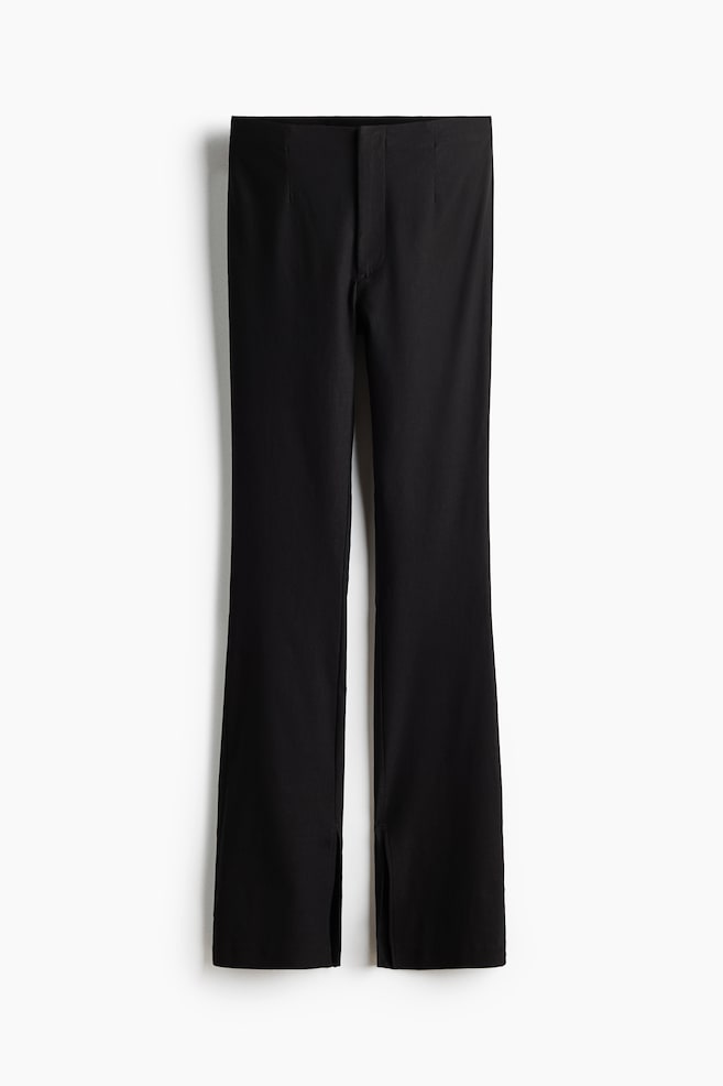 Black, high-waisted trousers are facing the front, displaying a slim-leg silhouette with vertical slits at the center of each hem. The trousers are featuring a clean waistband with a concealed front closure and subtle darting for a structured fit. - 1