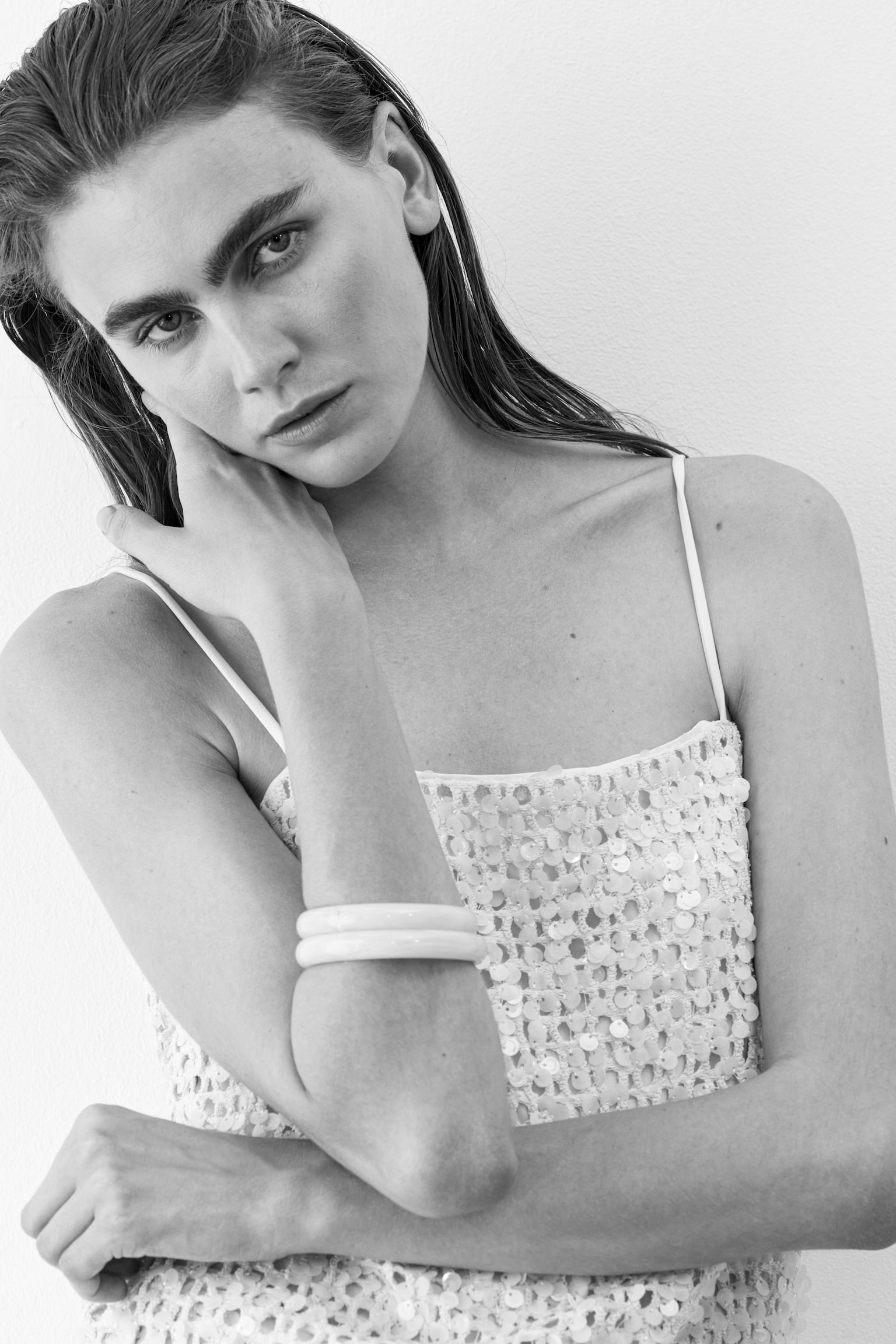 A white sequined top with thin spaghetti straps is worn, its fabric adorned with circular sequins. Two smooth, white bangles are stacked on the right forearm. - 1