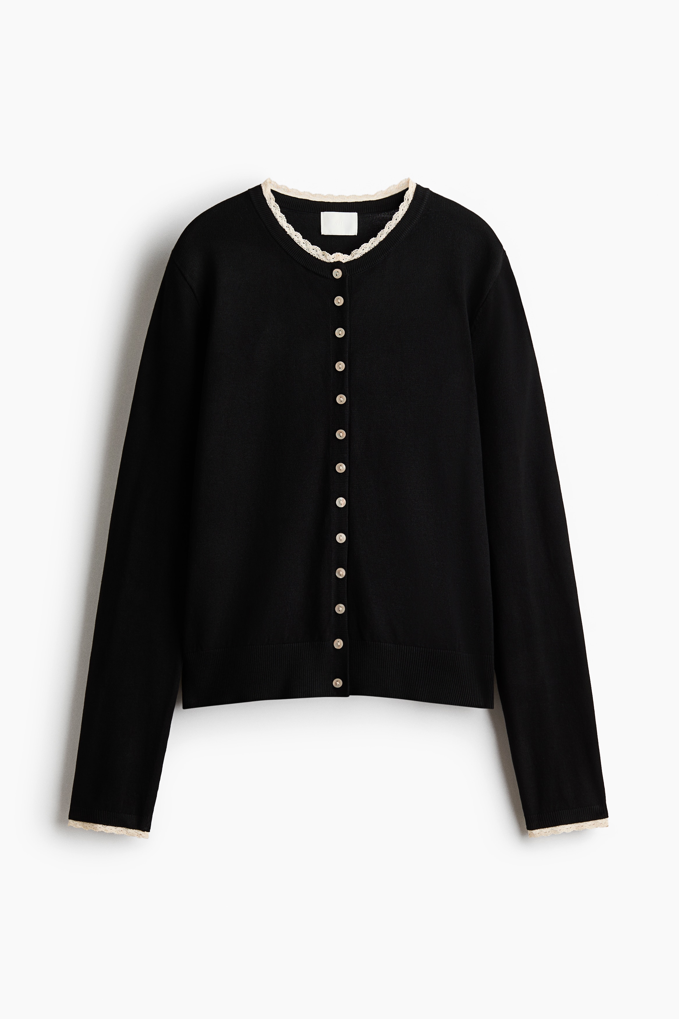 A black long-sleeve cardigan in a fine knit is shown from the front, featuring a round neckline with delicate cream lace trim and small buttons down the front. The slim-fit cardigan is styled partially unbuttoned and paired with matching black pants.