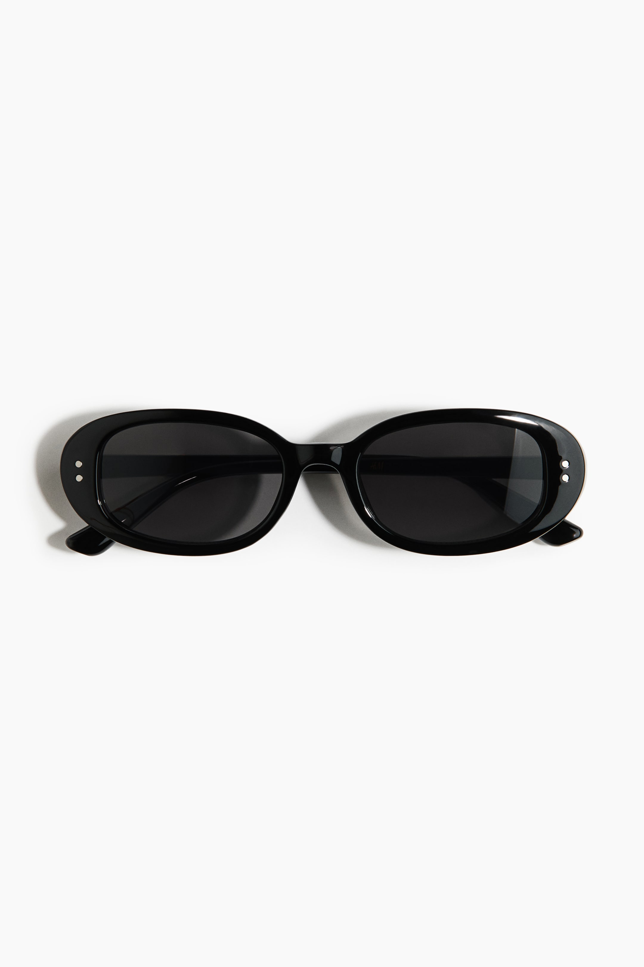 Ver imagen más grande: Se muestran unas gafas de sol ovaladas de frente, con monturas y varillas gruesas de plástico negro. Las lentes de plástico tintadas oscuras están encajadas en las monturas, con dos pequeños detalles de remache circular visibles en cada esquina exterior.