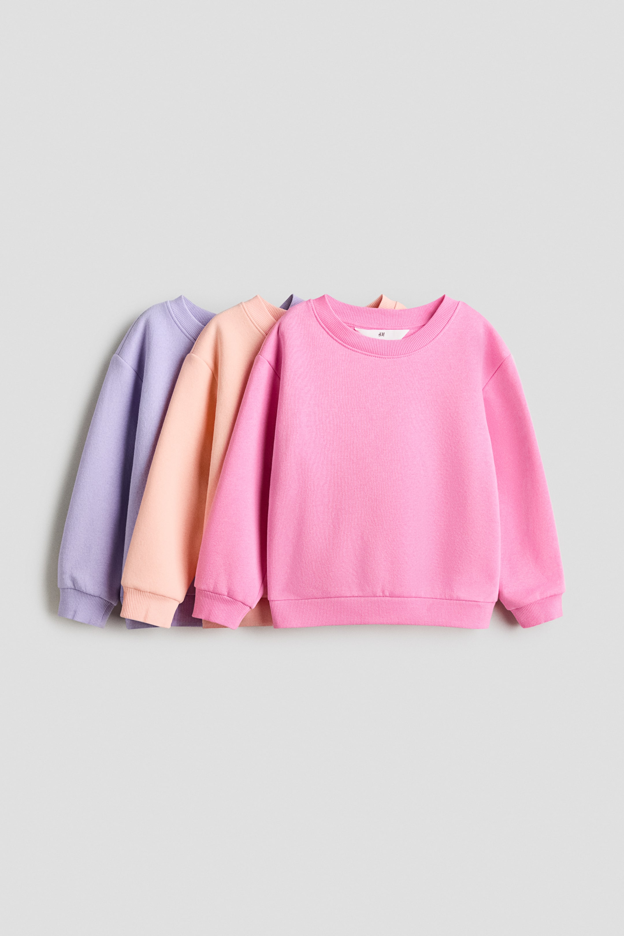 Grotere afbeelding bekijken: Drie tops met lange mouwen in sweatshirtstof zijn zichtbaar vanaf de VOORKANT, gepresenteerd in roze, perzik en lavendel. Elke top heeft een ronde halslijn, verlaagde schouders en ribboorden rond de halslijn, manchetten en zoom.