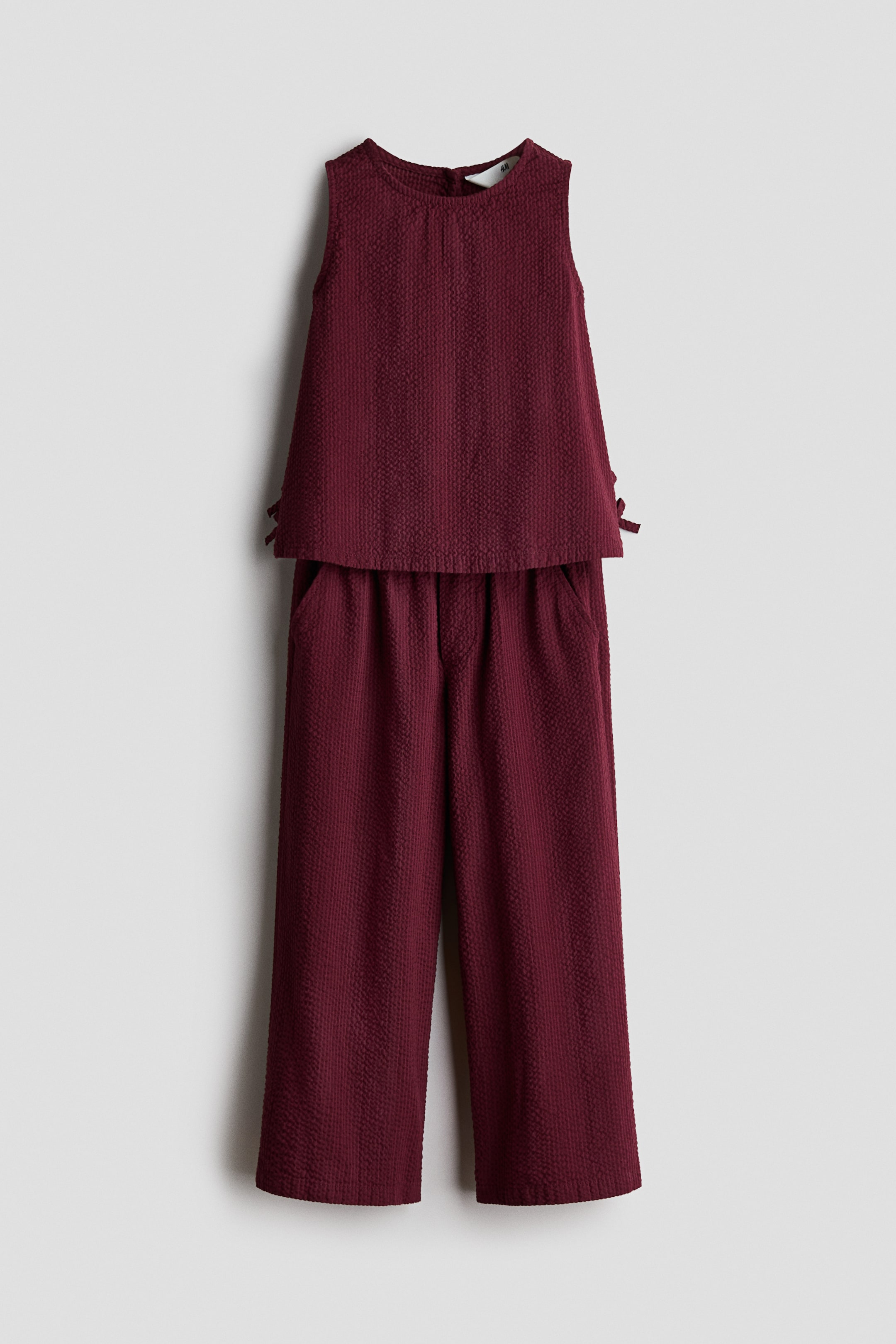 View larger image: This front view showcases a burgundy two-piece set made from crinkled, textured fabric. The sleeveless top features a round neckline and small decorative ties at the side hems, while the matching wide-leg trousers fall straight with visible side slip pockets.