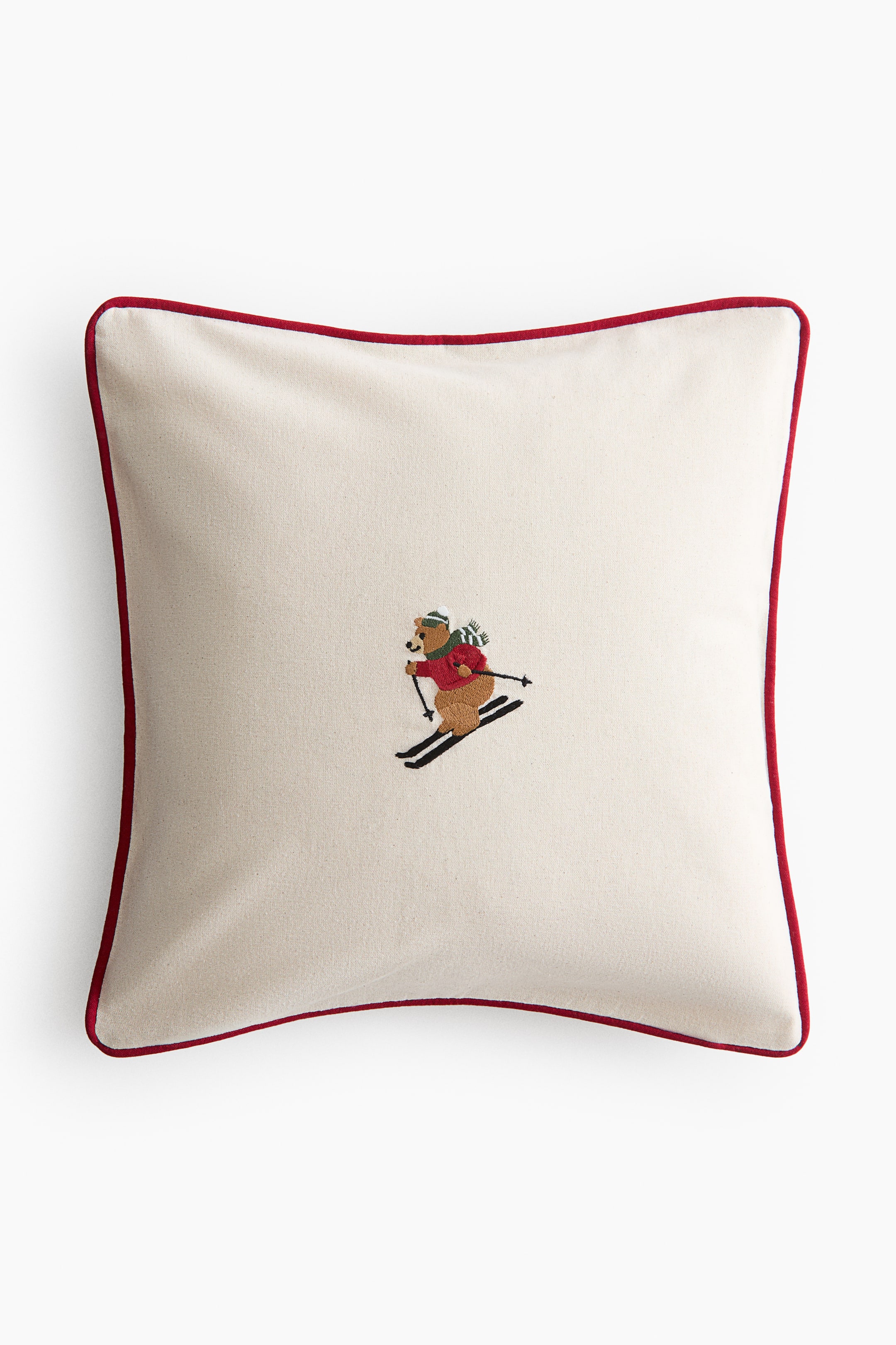 View larger image: A square cushion is shown from the front, featuring an off-white fabric with red piping along the edges. The centre of the cushion displays an embroidered brown bear, wearing a red sweater, a green and white striped scarf, and a green hat, and skiing with black skis and poles.