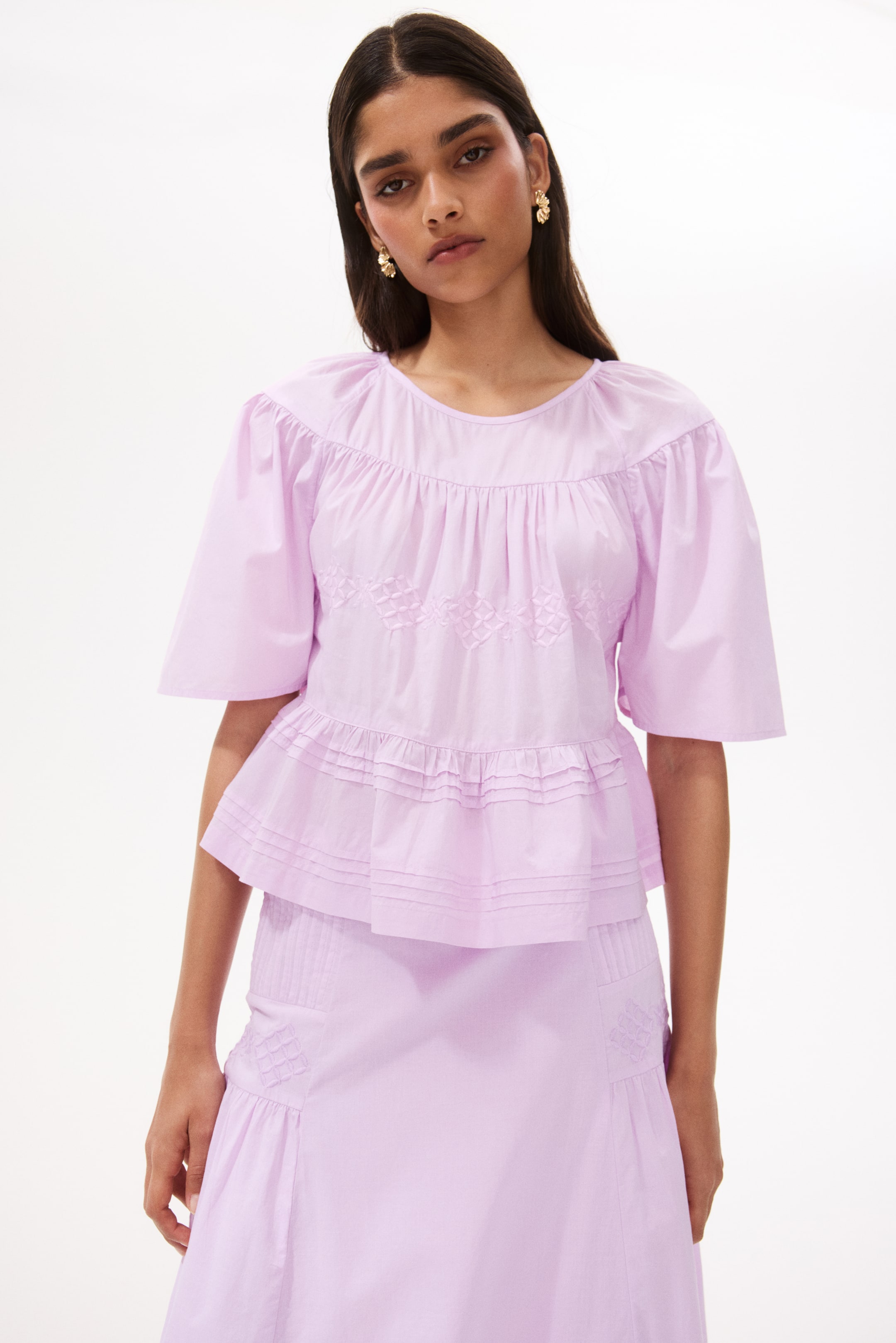 View larger image: A light purple top featuring a gathered round neckline and short voluminous sleeves. It showcases an embroidered geometric pattern across the chest, a tiered peplum with gathers, and horizontal pintucks along the bottom hem.