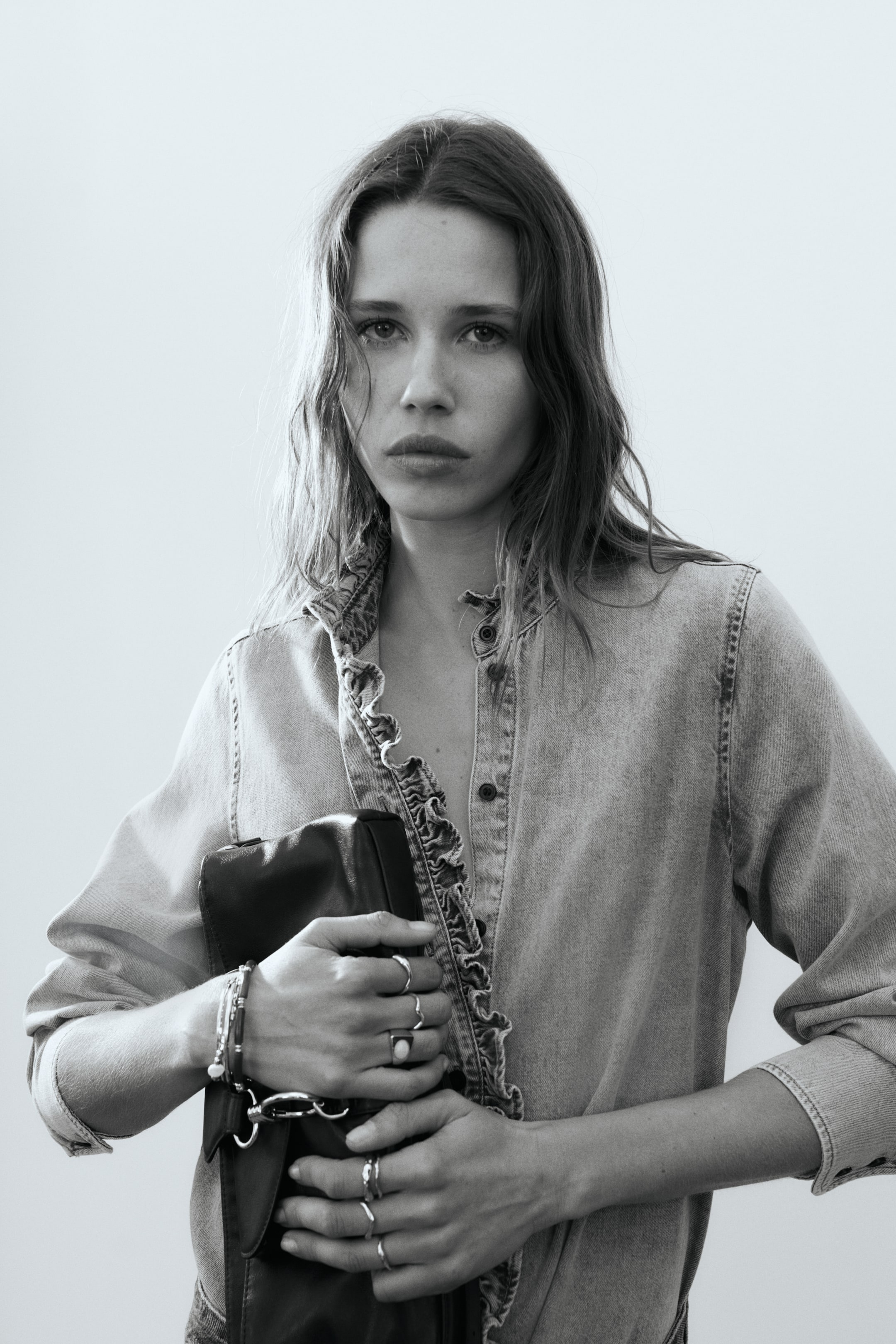 Zobacz większy obraz: Koszula z jasnego dżinsu jest noszona, z falbanami wzdłuż przedniego zapięcia i stójką. Koszula jest częściowo rozpięta, a jej długie rękawy są widoczne, gdy jest prezentowana z lekko przedniego kąta.