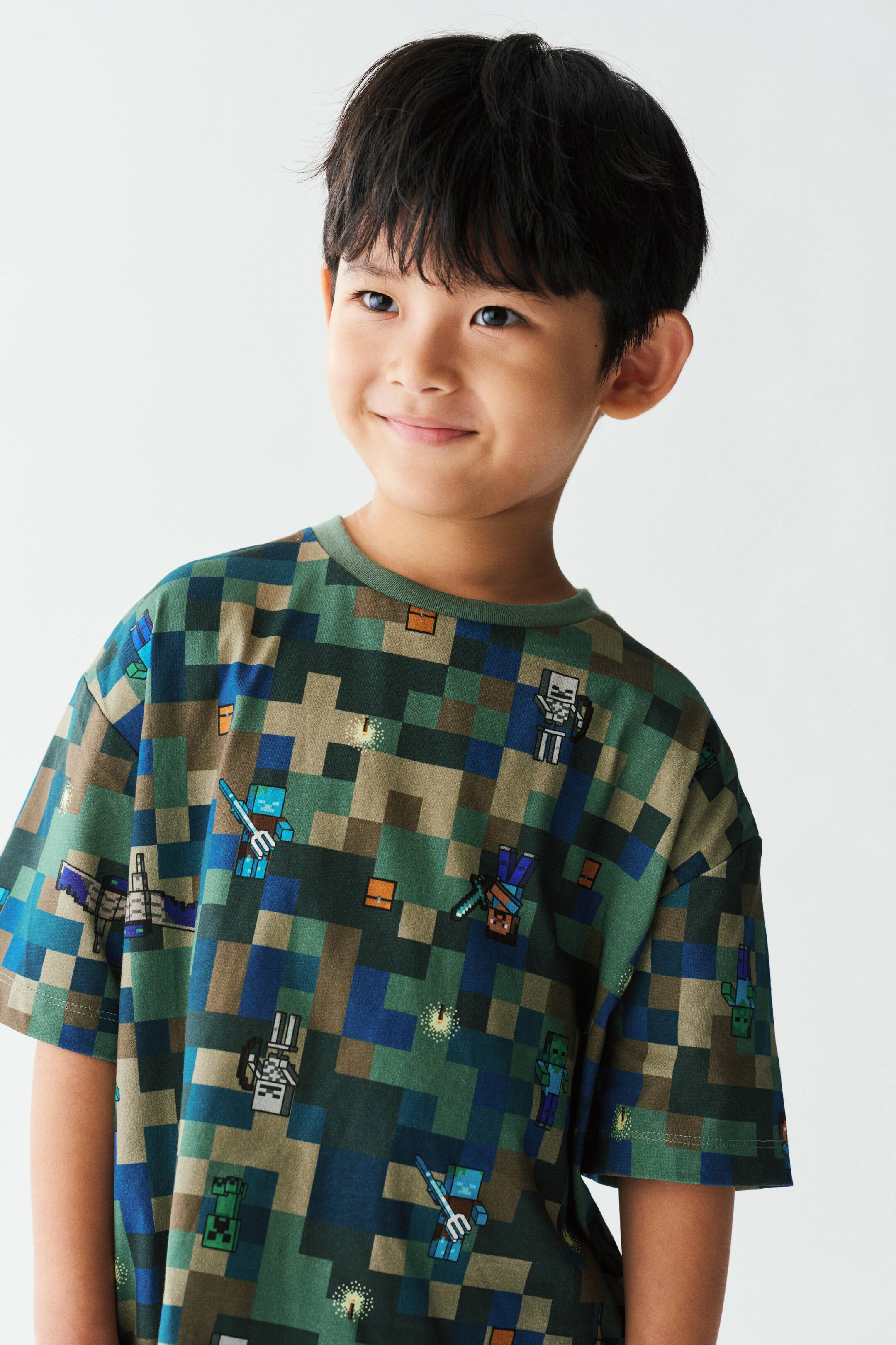 View larger image: A green and blue pixelated print tee in soft cotton jersey is being worn by the model. The tee features a round, rib-trimmed neckline, dropped shoulders, and a printed design of blocky video game characters.