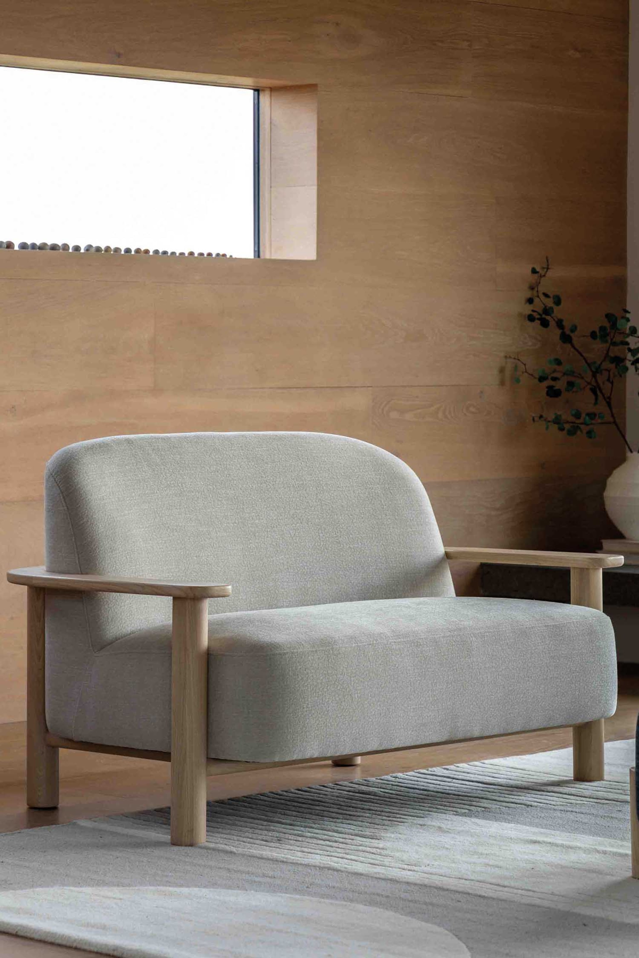 View larger image: A two-seater sofa featuring light grey textured fabric upholstery, a gently curved backrest, and a single seat cushion. It is structured with a light wooden frame, including flat, rounded armrests and stout wooden legs, and is facing slightly to the right.