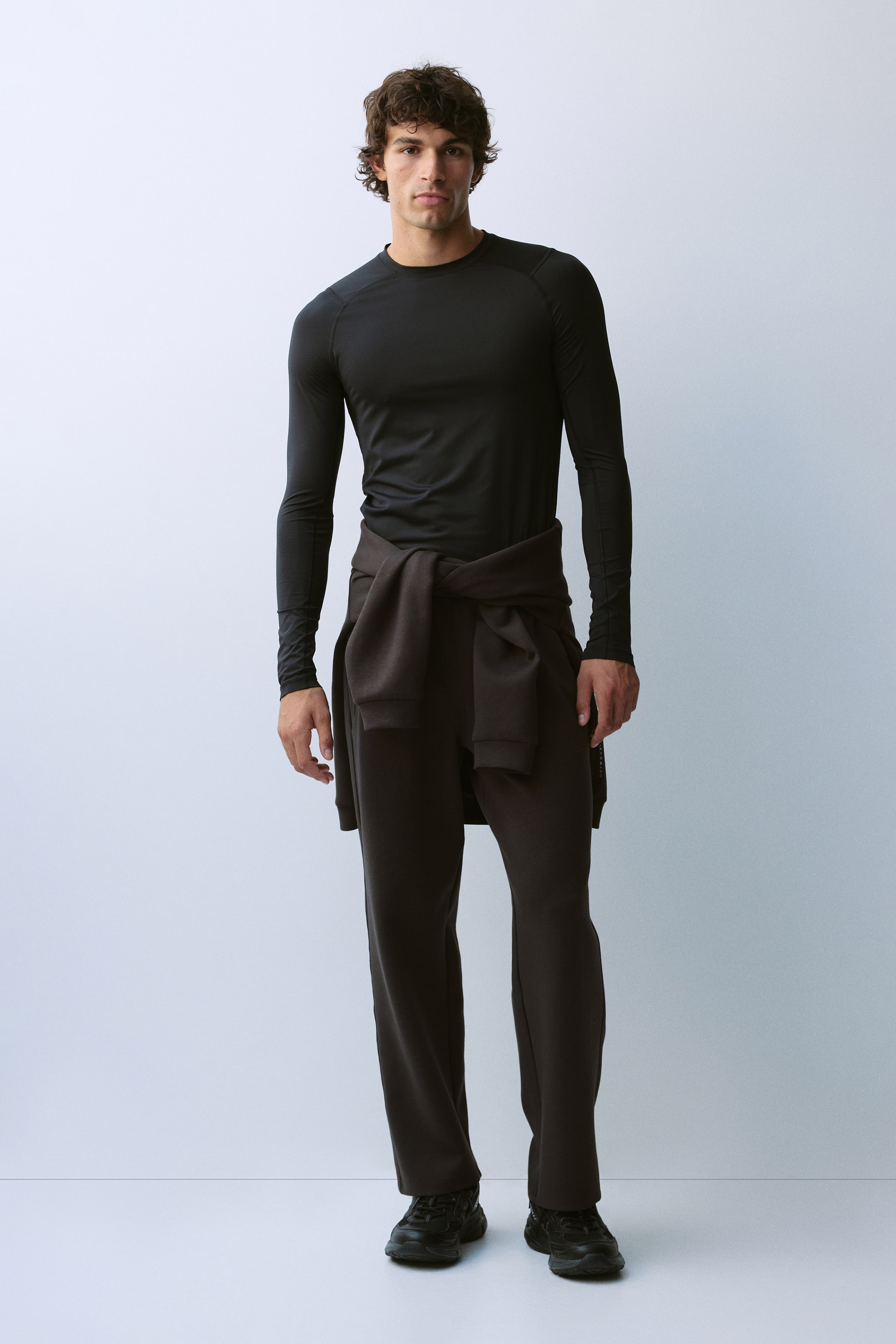 View larger image: The model is wearing a black, fitted long-sleeve athletic top with a crew neck and raglan seams. The top is styled with dark brown wide-leg sweatpants and a matching sweatshirt tied around the waist.