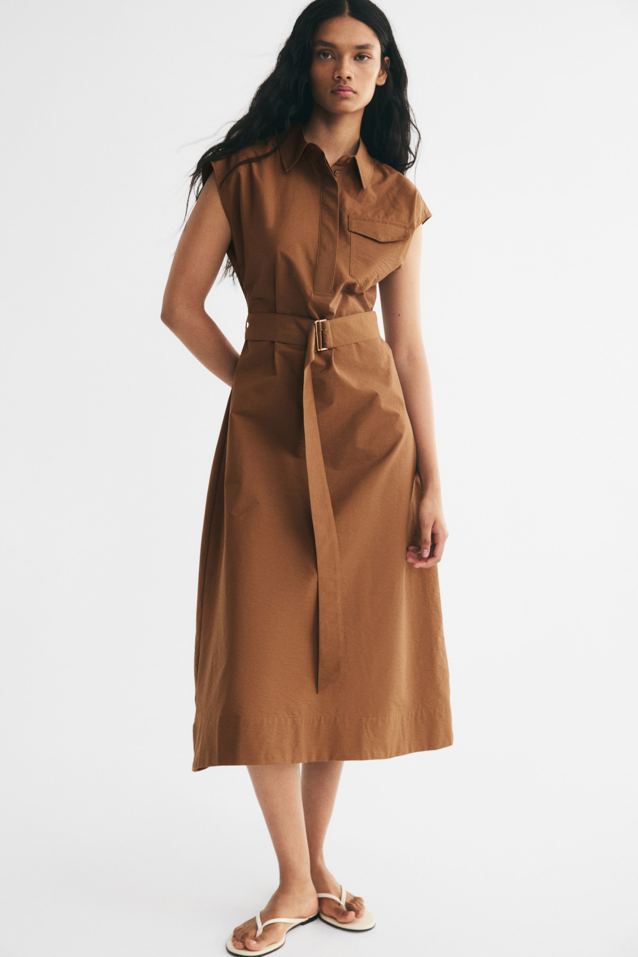 View larger image: A brown calf-length, sleeveless dress featuring a collar, concealed button placket, flap chest pocket, and wide waist belt with a metal D-ring buckle is being worn by the model. Its gently flared skirt falls to a straight hem, and white flip-flops are on the model’s feet.