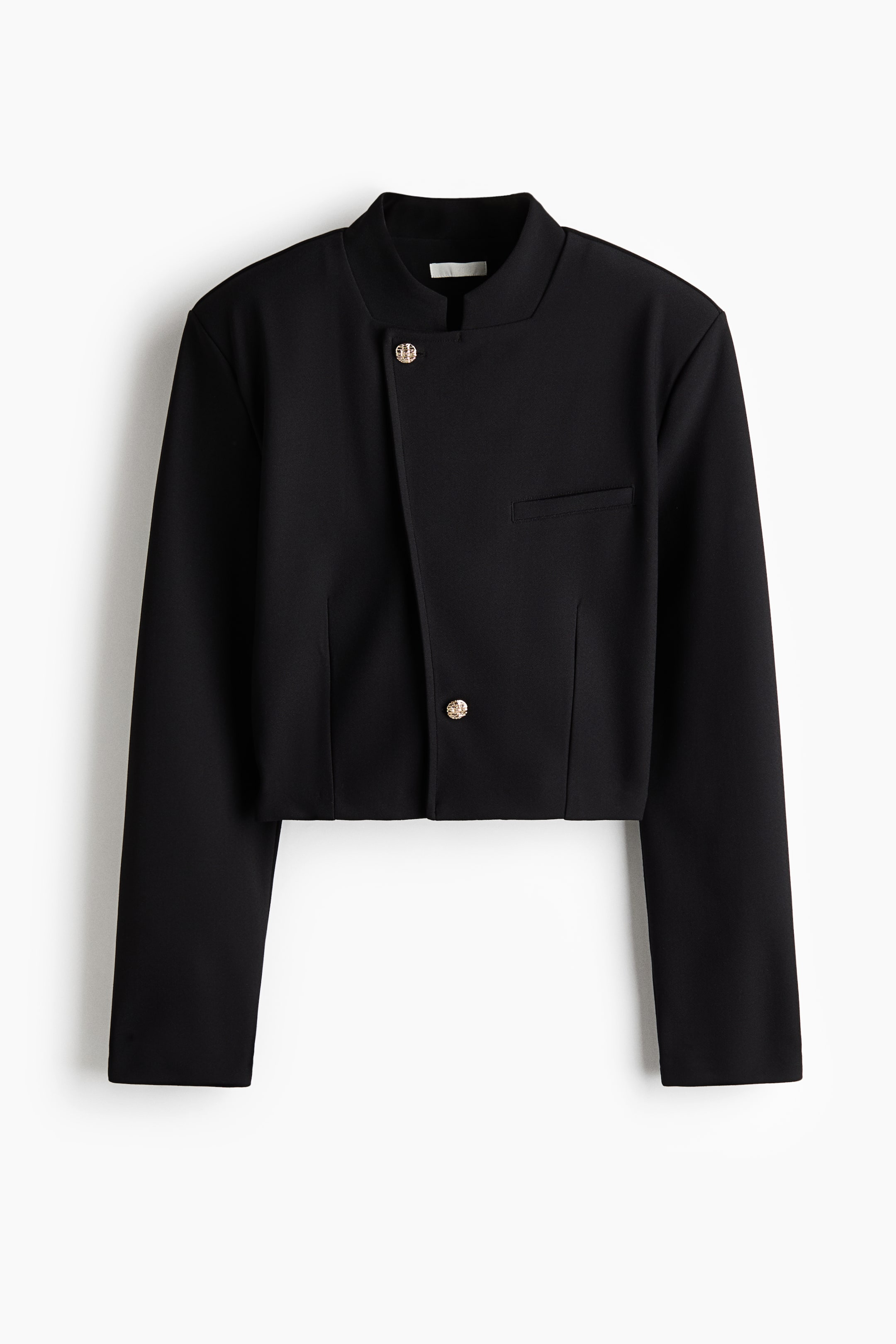 View larger image: A black cropped jacket is shown from the front, featuring a structured band collar and an asymmetrical front closure secured with two textured gold-tone buttons. The jacket has a single welt pocket on the left chest and long, straight sleeves.