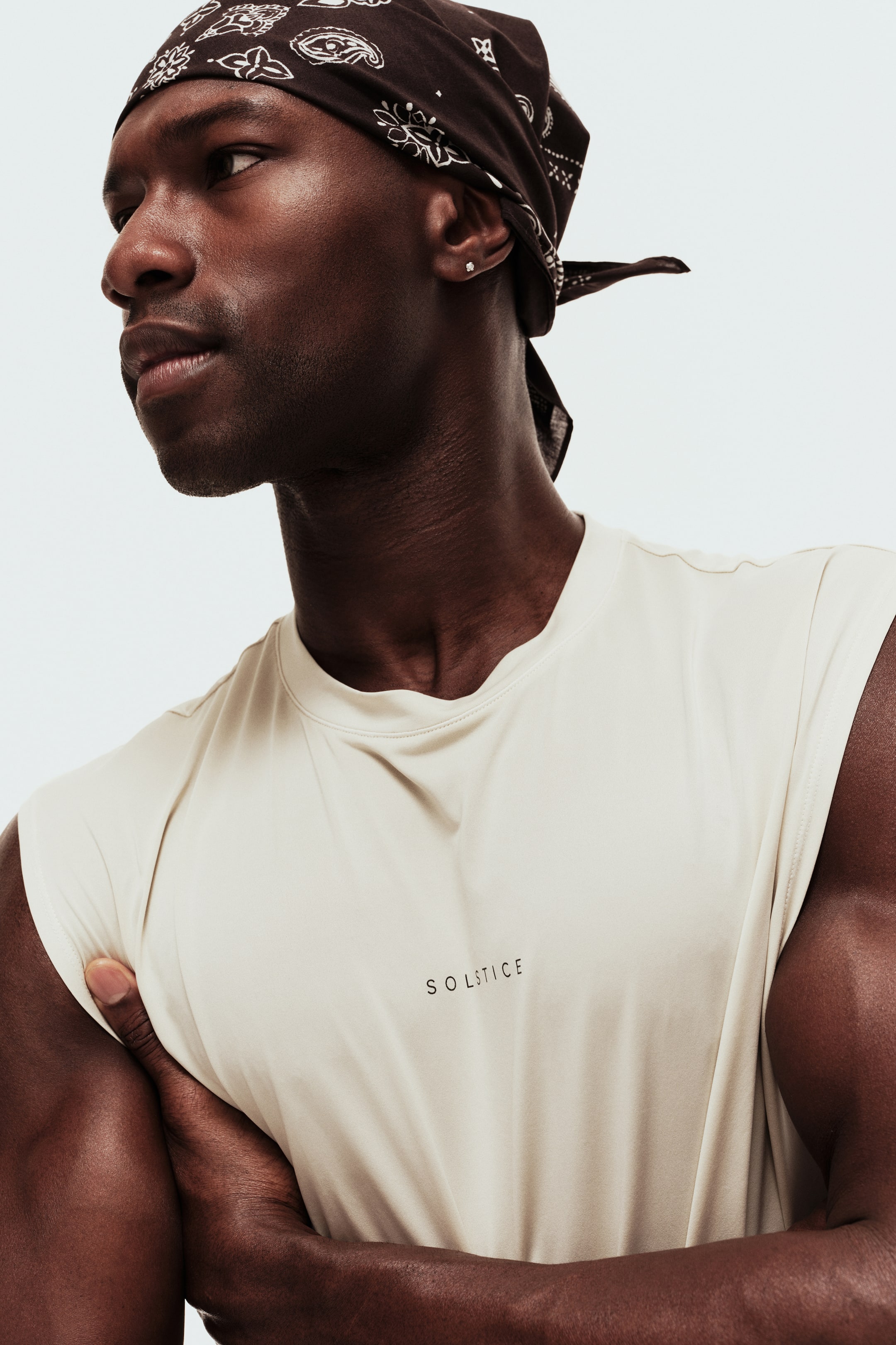 View larger image: A light beige sleeveless top with a crew neck and "SOLSTICE" text is worn by the model. The model has their arms crossed over their chest, subtly pulling the fabric of the top.