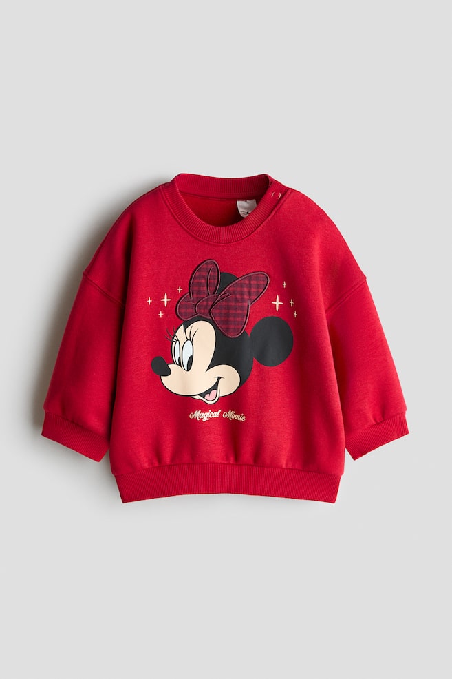 A red toddler sweatshirt is lying flat, showcasing a front-facing design with a Minnie Mouse graphic wearing a textured red and black plaid bow. The sweatshirt features a ribbed crew neck with shoulder snap buttons, dropped shoulders, ribbed cuffs, and a ribbed hem. - 1