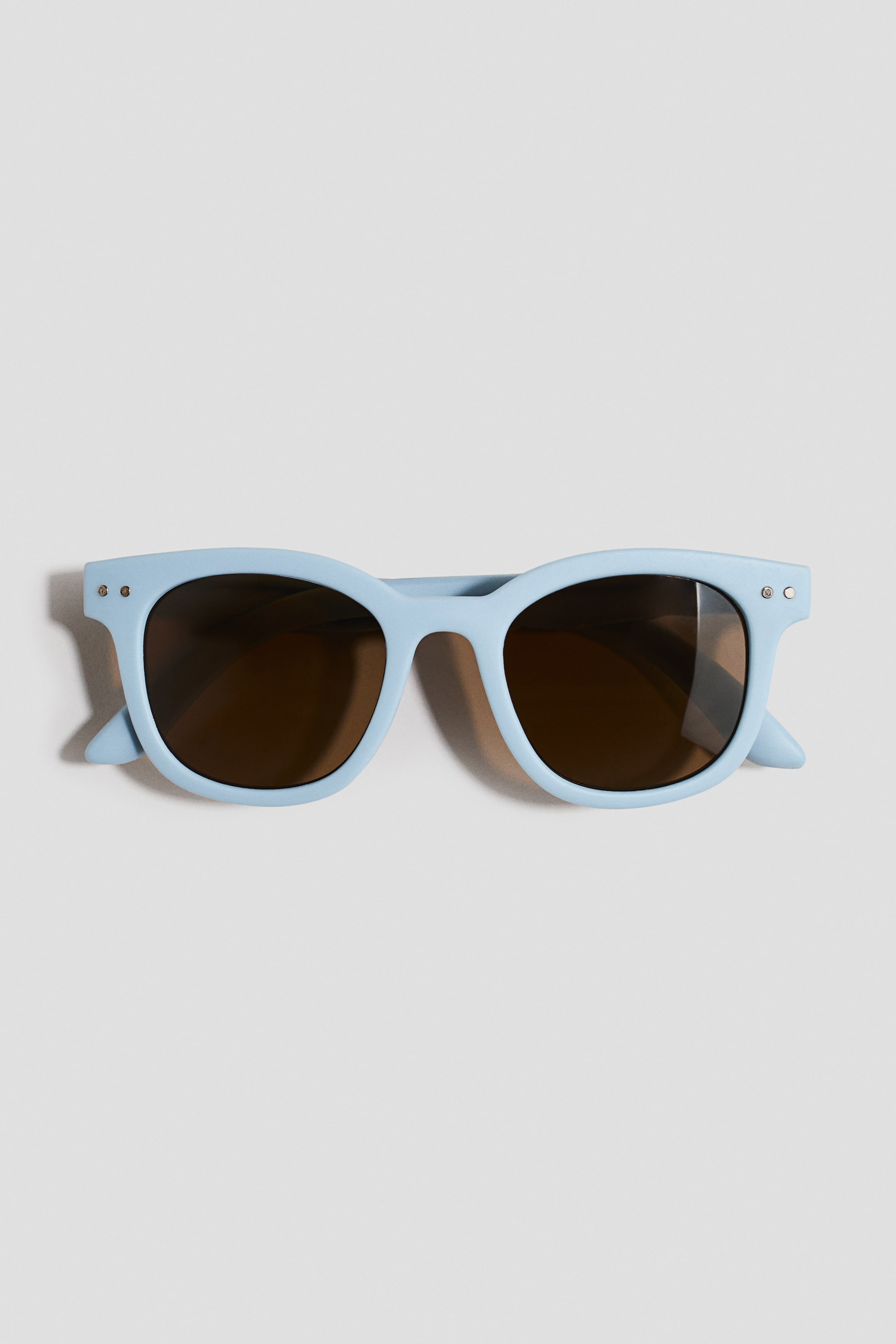 View larger image: Light blue sunglasses are laid flat, showcasing a rectangular frame with rounded edges, dark brown lenses, and small silver rivets visible on the front upper corners.