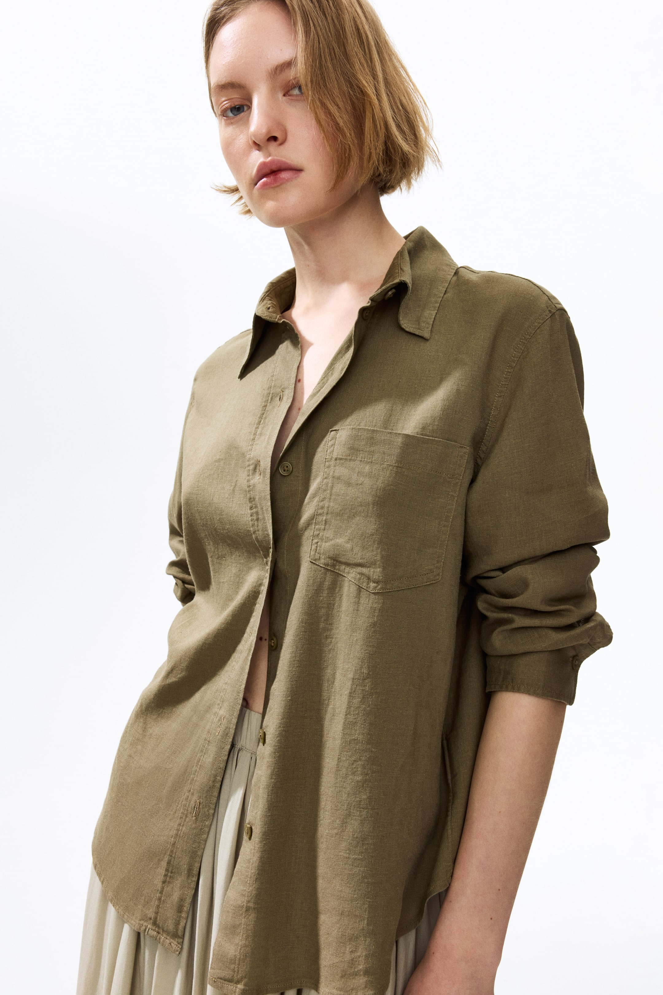 View larger image: The model is donning a green shirt crafted in a breezy weave, complete with a collar and an open chest pocket. The shirt's front buttons are partially undone, and its long sleeves with buttoned cuffs are casually rolled up. The rounded hem elegantly falls over light-hued trousers.