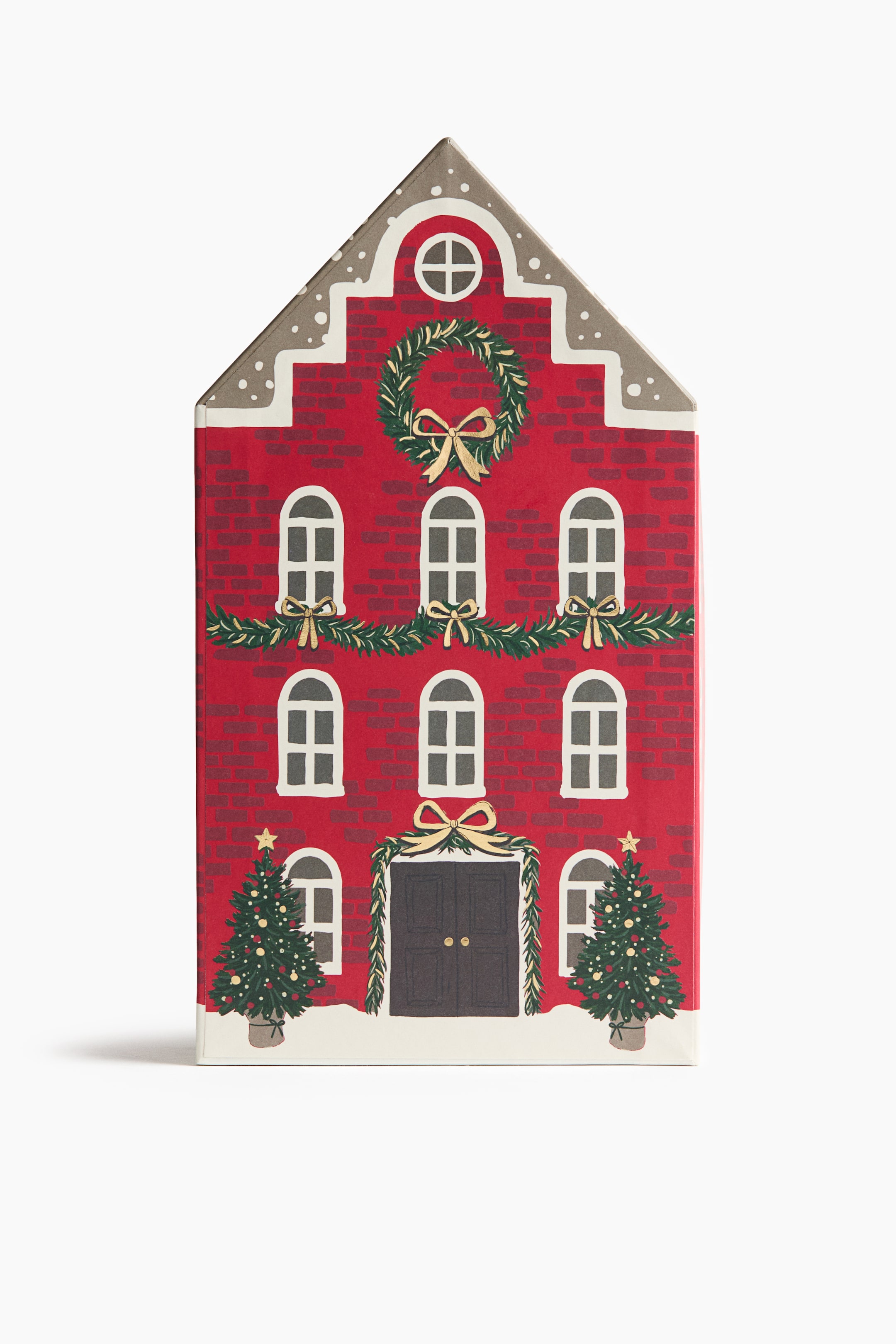 View larger image: A house-shaped Advent calendar is standing upright, showcasing a red brick façade designed to resemble a festive townhouse from the front. The detailed illustration features rows of arched windows, a dark double door, green garlands with gold bows, a large holiday wreath, and two decorated Christmas trees flanking the entrance at the snowy base.