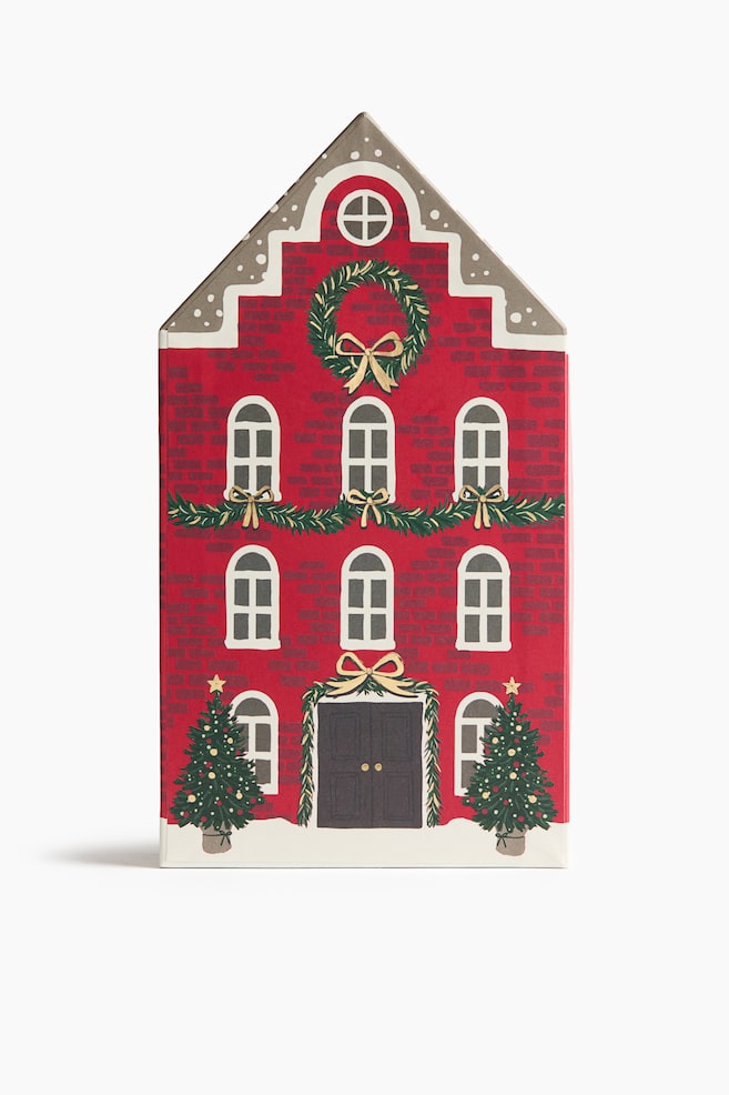 A house-shaped Advent calendar is standing upright, showcasing a red brick faÃ§ade designed to resemble a festive townhouse from the front. The detailed illustration features rows of arched windows, a dark double door, green garlands with gold bows, a large holiday wreath, and two decorated Christmas trees flanking the entrance at the snowy base. - 1