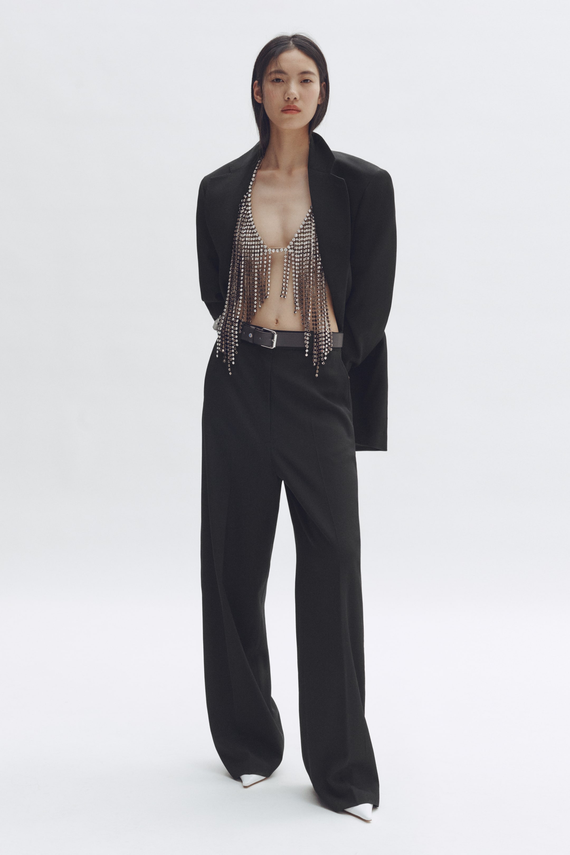 View larger image: A black blazer drapes open over a silver chainmail bralette, adorned with numerous dangling, reflective strands. Black wide-leg trousers are paired with a black belt, while white pointed-toe shoes peek from beneath the trousers.