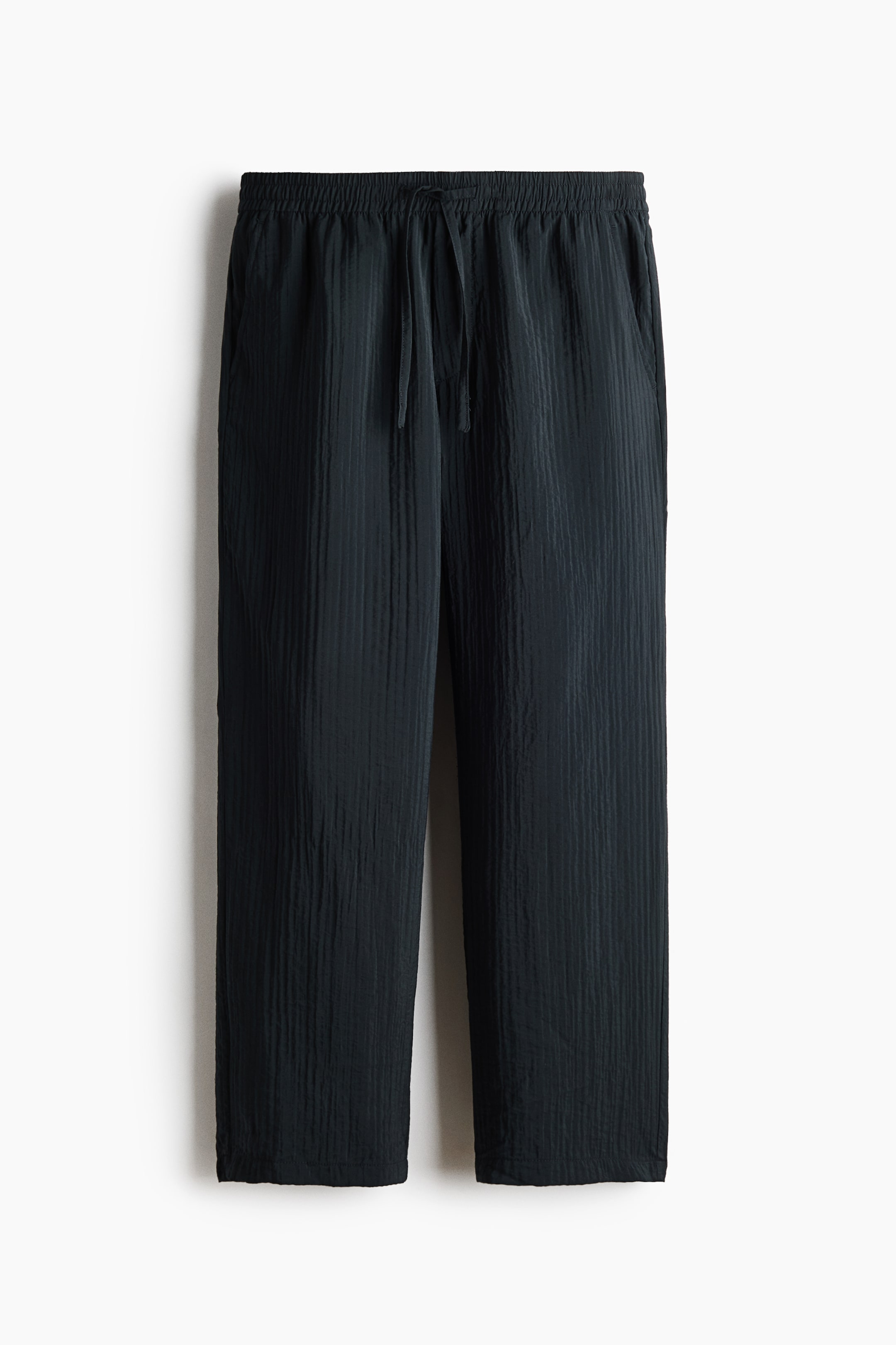 View larger image: The dark grey pants are shown from the front, featuring a crinkled, vertically textured fabric, an elasticated waistband with a drawstring tie, side pockets, and a relaxed straight-leg fit.
