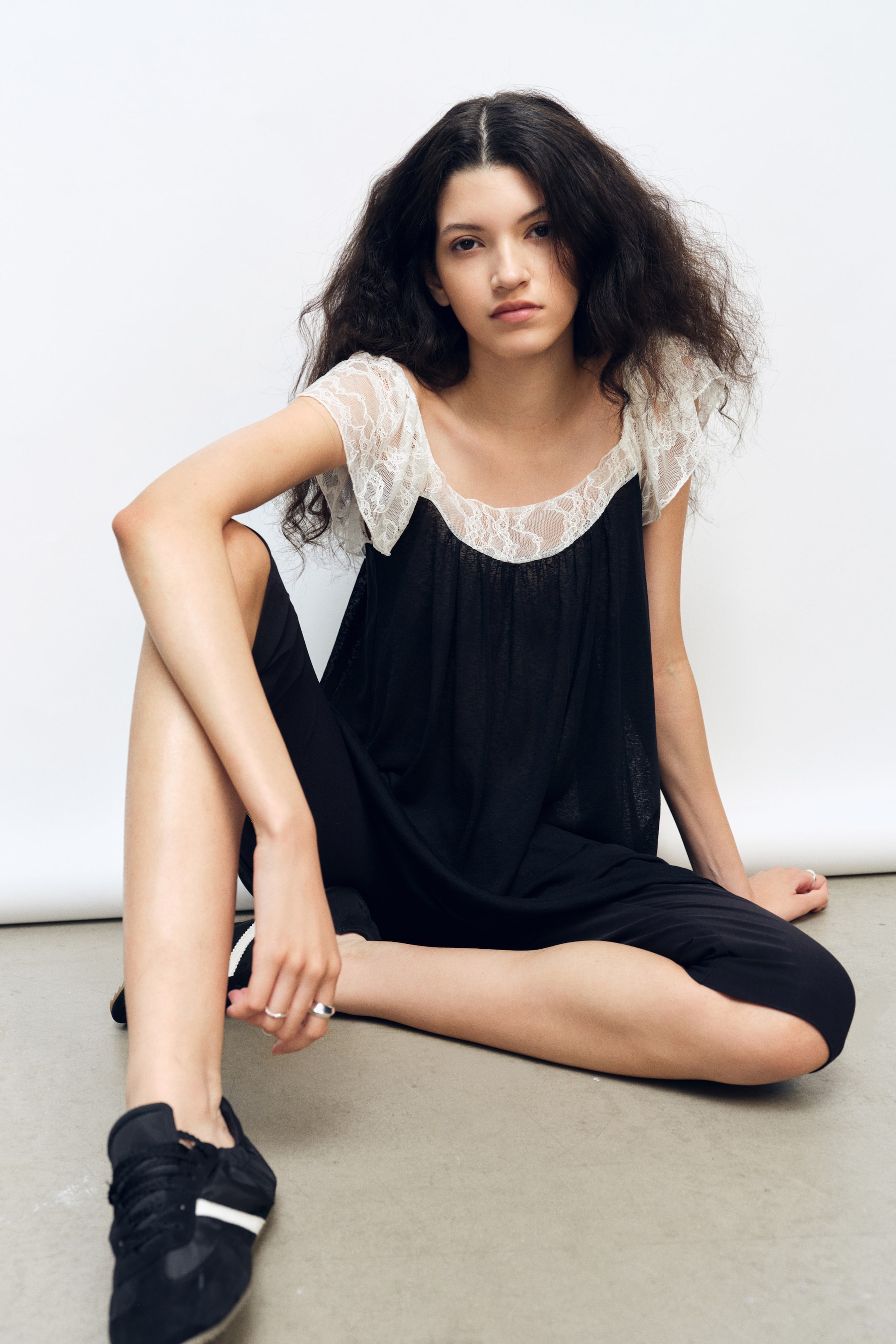 Größeres Bild anzeigen: Ein locker sitzendes schwarzes Mini-Kleid wird von dem Model getragen, das auf dem Boden sitzt. Das Kleid verfügt über einen gerafften Rundhalsausschnitt mit kontrastierendem weißen Spitzen-Einsatz und dazu passenden flatternden Spitzenärmeln.