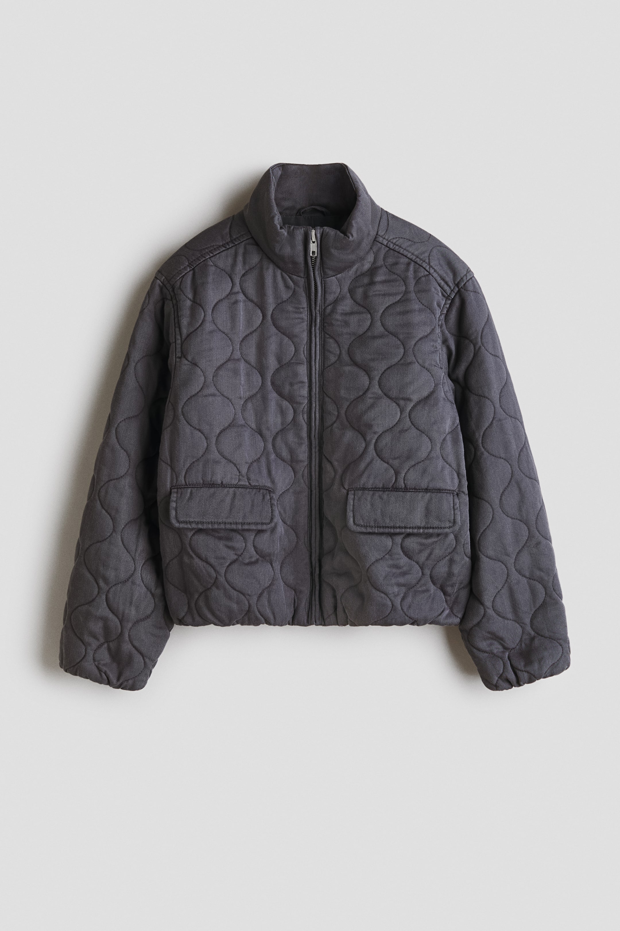 View larger image: A charcoal quilted jacket is showcased in a front view, highlighting an organic wavy pattern. It features a full zipper closure, a stand-up collar, two rectangular front patch pockets, and an elasticized bottom hem.