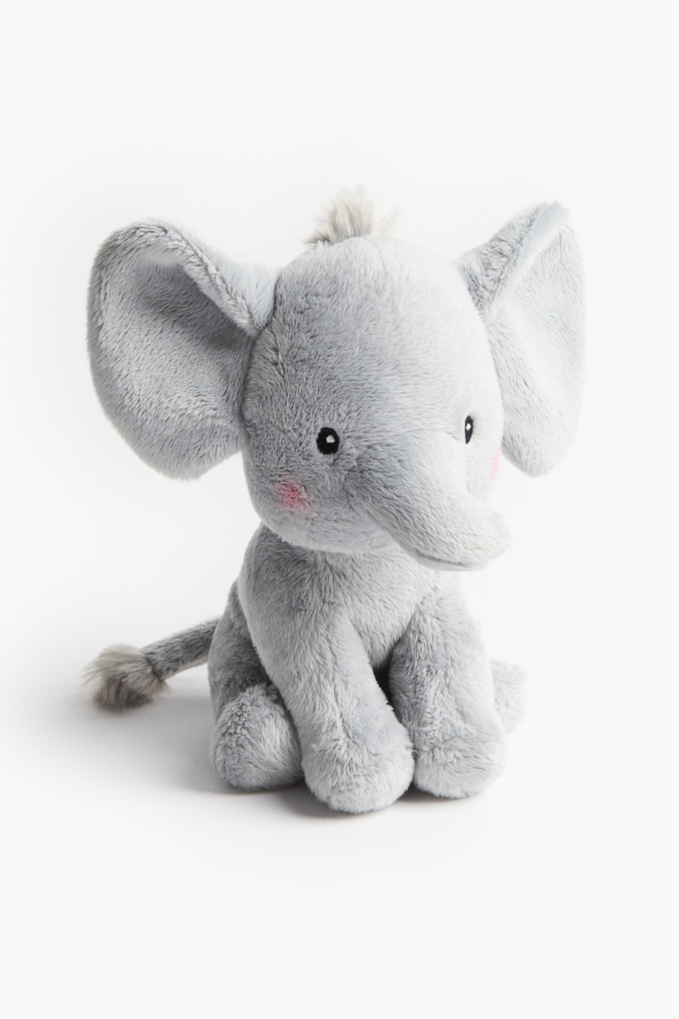 View larger image: A soft toy elephant is sitting, facing front, and made of grey plush material. It features large floppy ears, a tuft of grey fur on its head, small black eyes, pink blush marks on its cheeks, a short trunk, and a small tail with a tuft of fur.