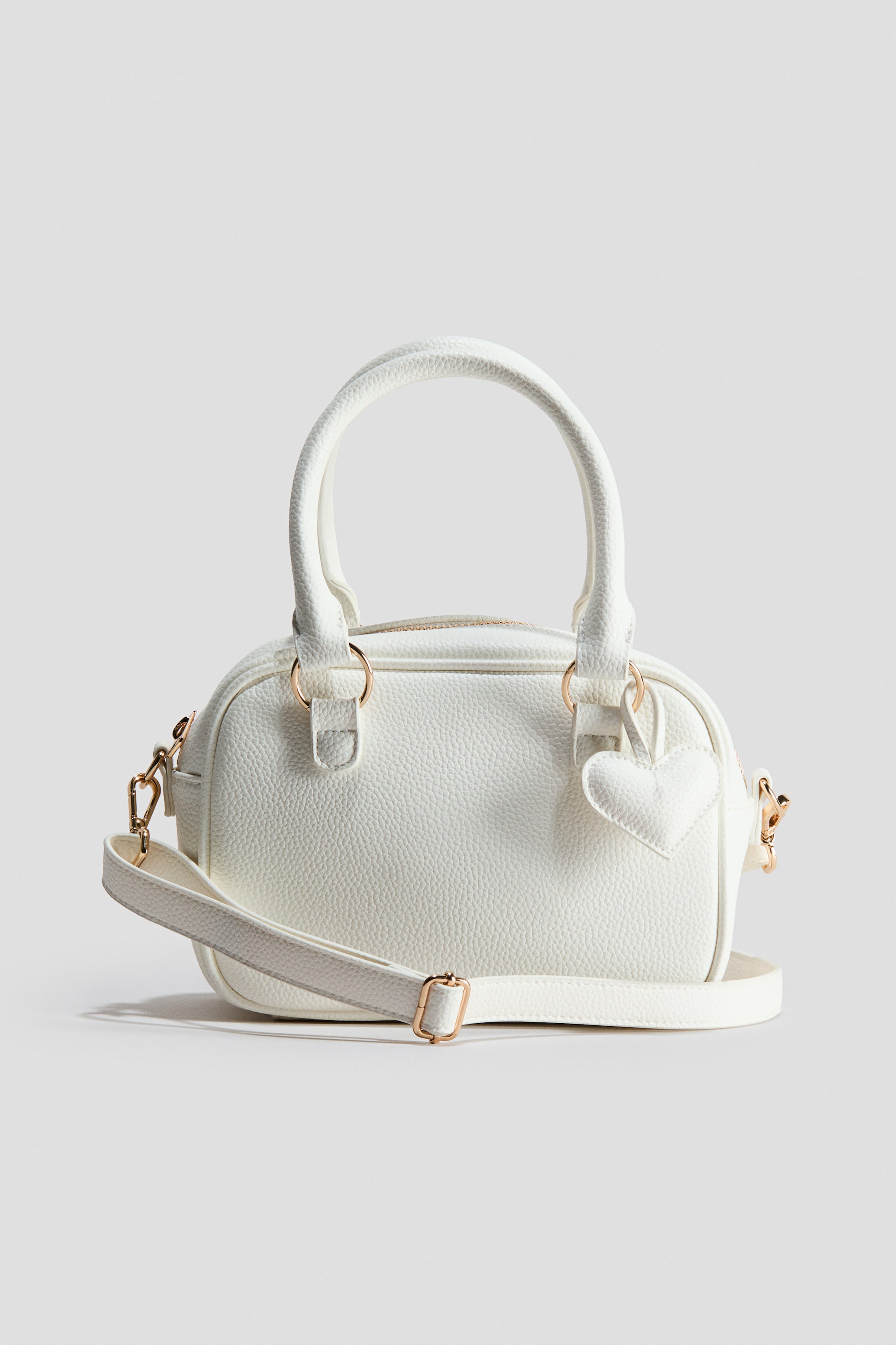 Ver imagen más grande: Un pequeño bolso de hombro blanco en tejido recubierto texturizado está de frente, mostrando dos asas superiores y un cierre de cremallera en la parte superior. Un charm en forma de corazón cuelga de una de las uniones de las asas, y una correa de hombro desmontable y ajustable con herrajes en tono dorado descansa sobre su frente.