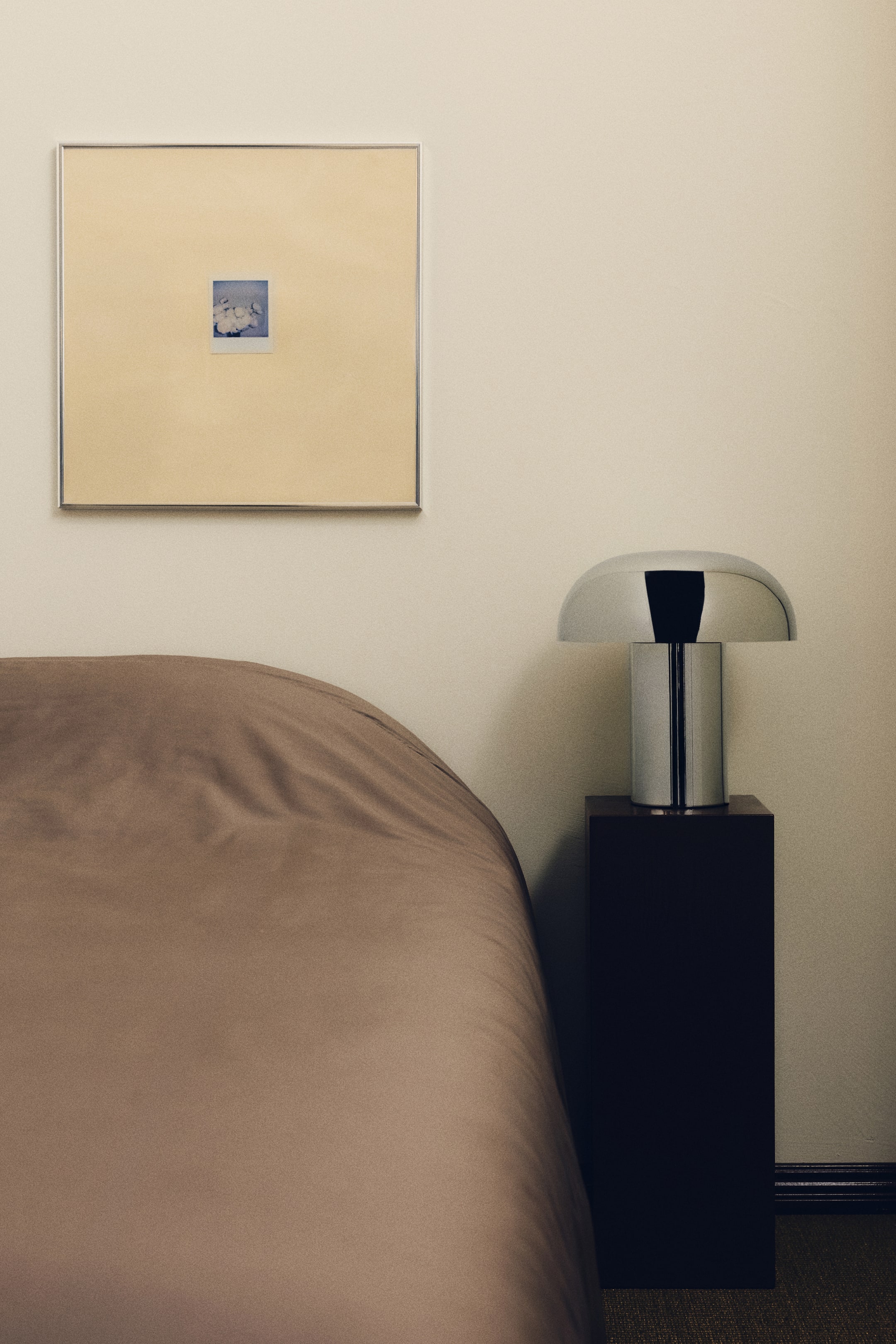 View larger image: A polished metallic table lamp features a rounded, mushroom-like shade and a cylindrical base. It rests on a dark rectangular pedestal, positioned with its front facing slightly to the left.