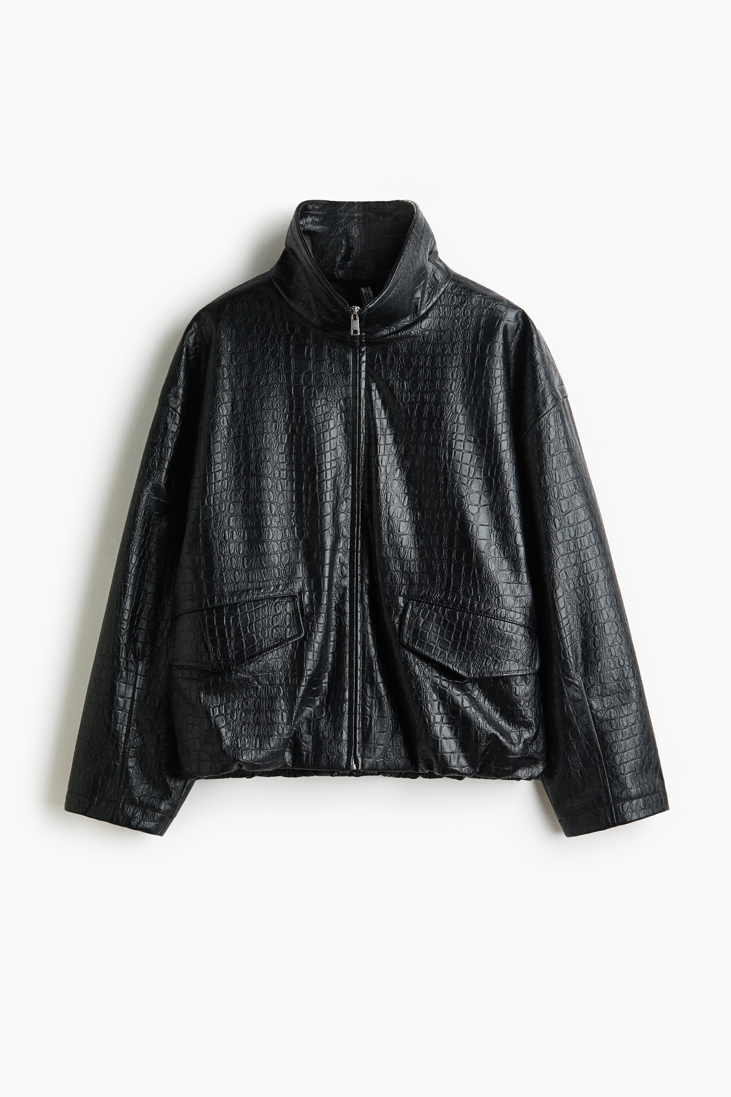 Eine oversized schwarze Jacke aus beschichtetem Stoff mit Krokodilstruktur wird vom Model Ã¼ber einem schwarzen Oberteil getragen. Das Model hÃ¤lt den Rand des Stehkragens mit einer Hand, wÃ¤hrend die andere Hand auf der HÃ¼fte ruht, wodurch die Ã¼berschnittenen Schultern und der FrontreiÃverschluss der Jacke betont werden.