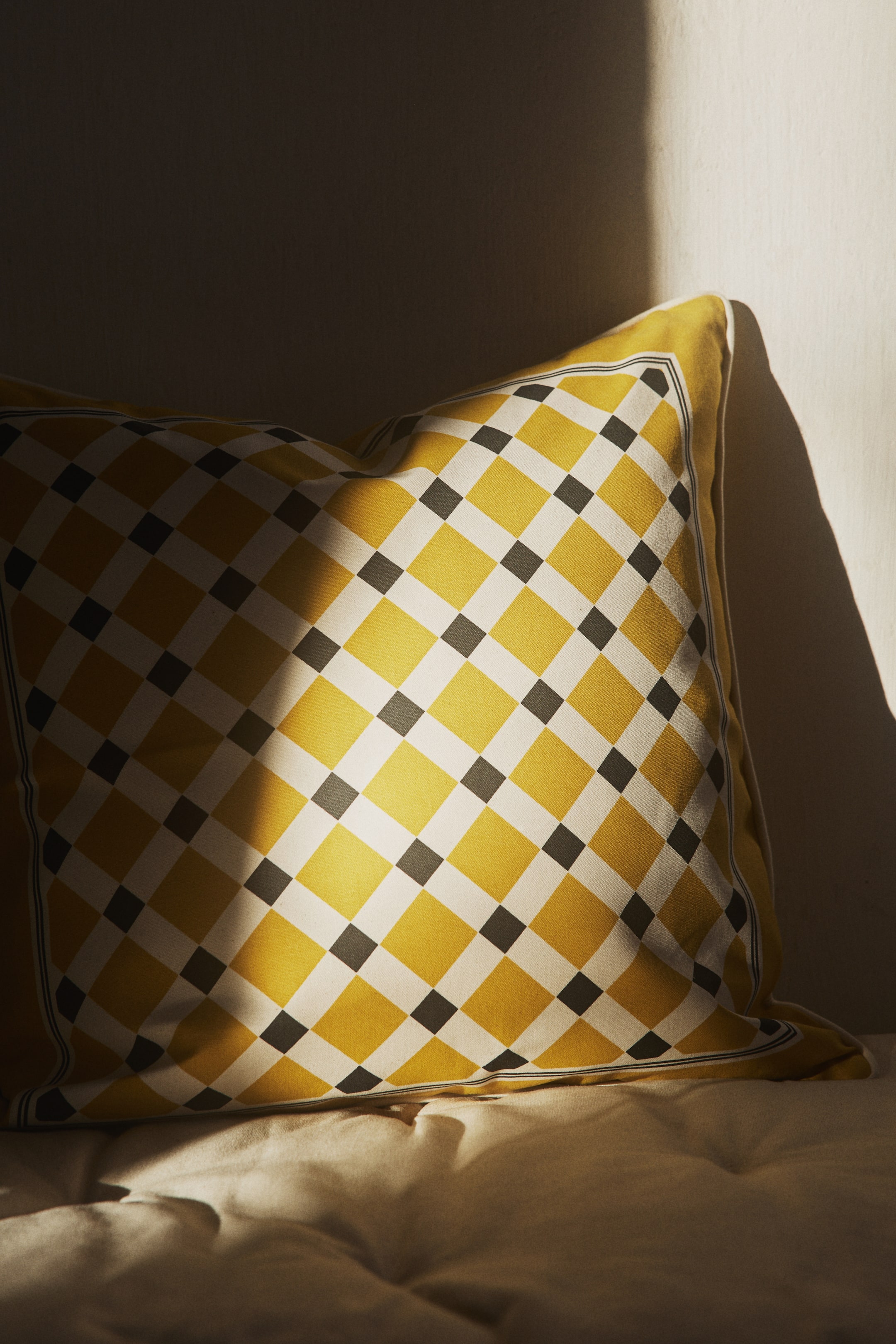 View larger image: The cushion cover is oriented frontally, showcasing a comprehensive harlequin design featuring yellow and off-white diamond shapes, highlighted by subtle black squares at the junctions. The visible edges are elegantly detailed with piping.