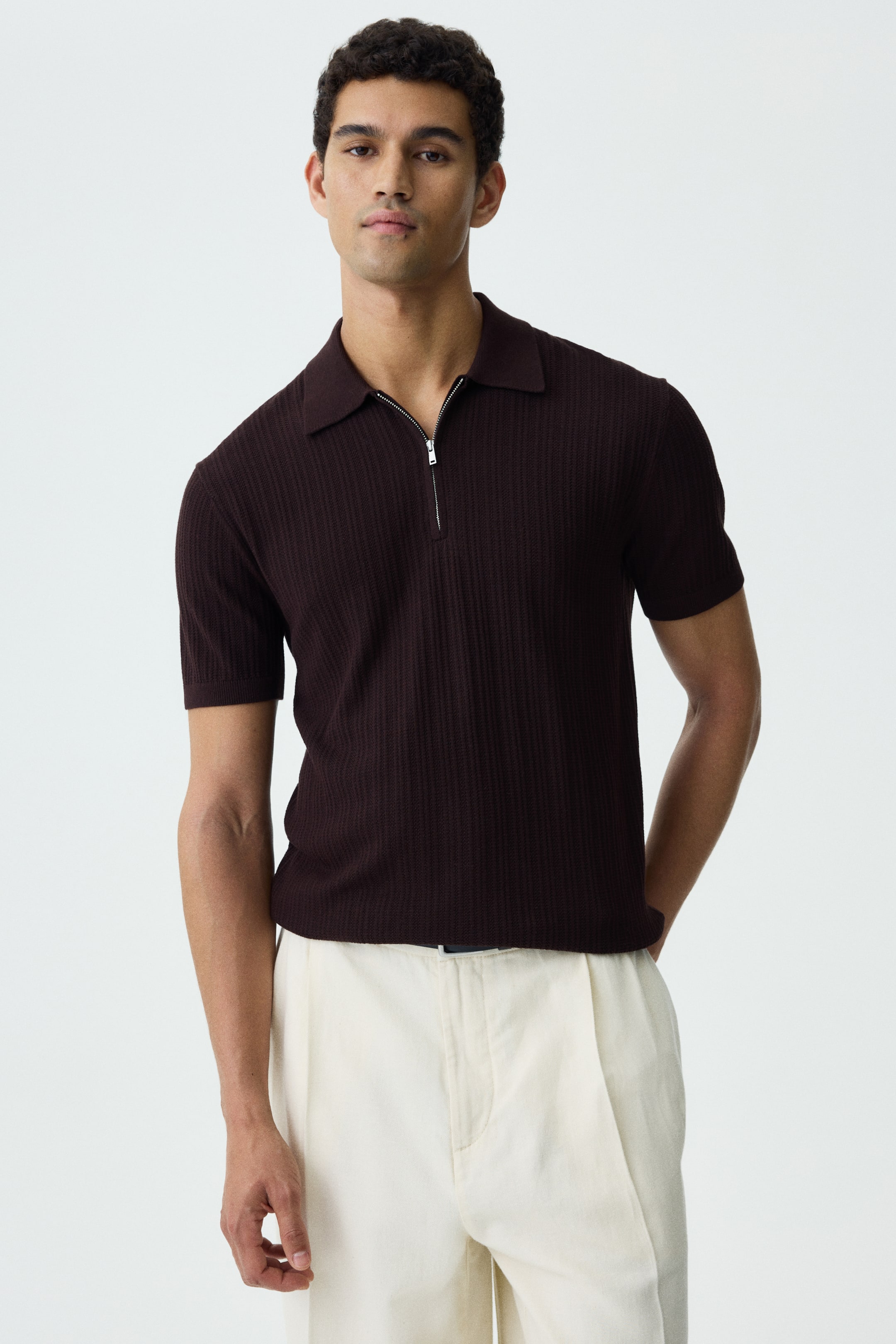View larger image: A dark brown textured knit polo shirt featuring a collar, a half-zip closure, short sleeves with ribbed cuffs, and a ribbed hem. The model is sporting the slim-fit shirt, neatly tucked into light-coloured trousers.