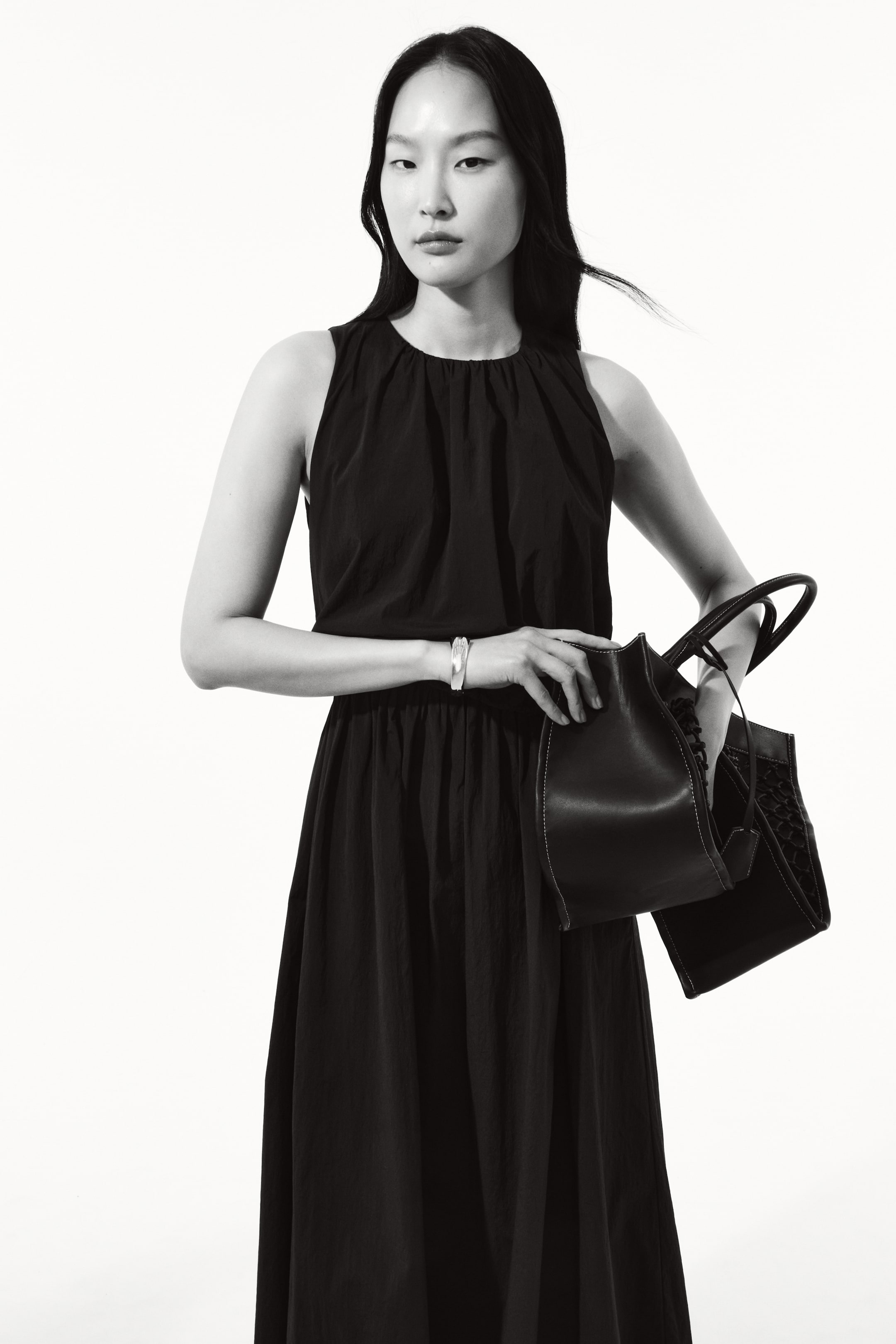 View larger image: The model is showcasing a black, sleeveless dress that falls to calf-length, featuring a gathered round neckline and a voluminous bodice. The dress is designed with a gathered seam at the waist. Accompanying the look, the model carries a sleek black handbag.