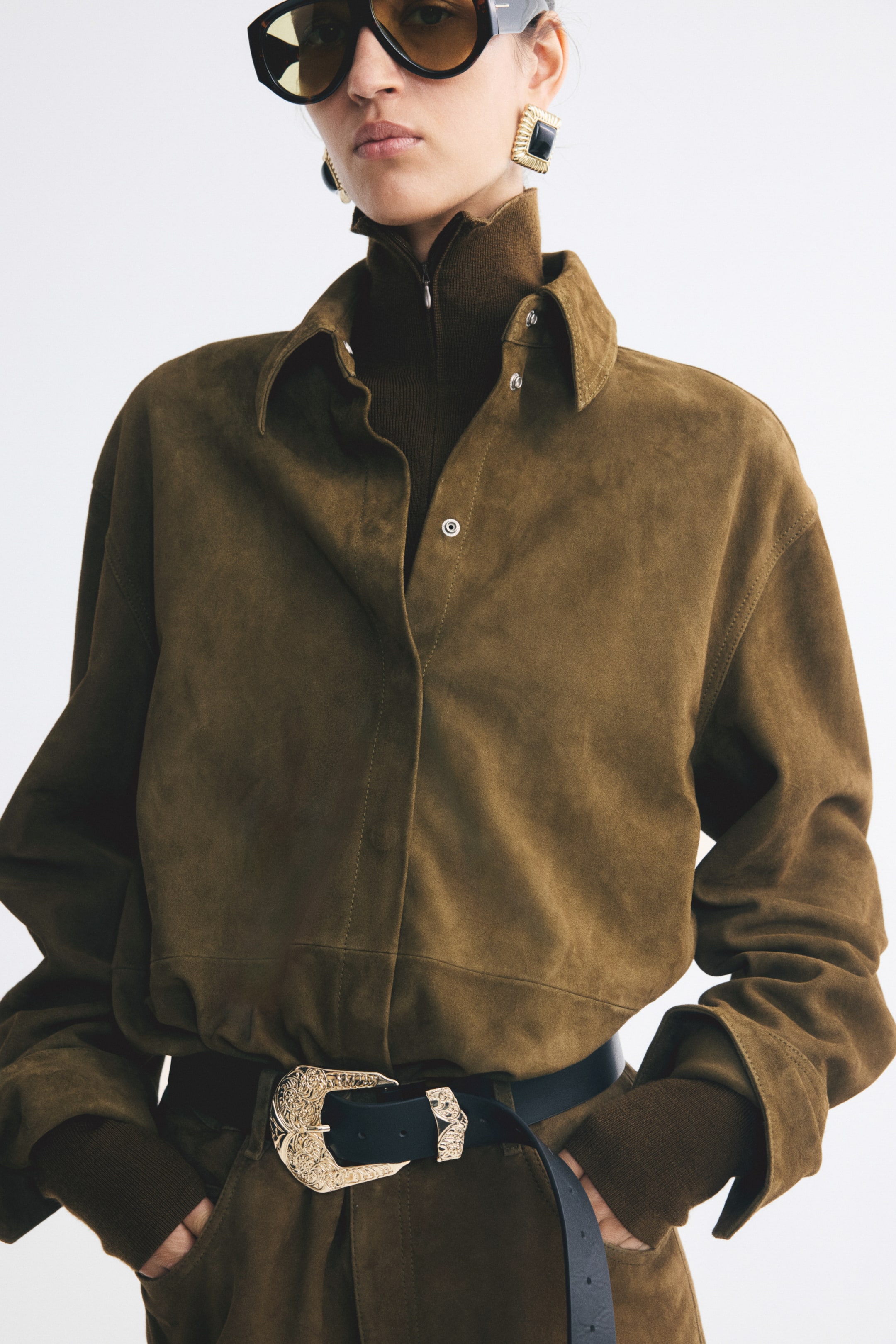 View larger image: An oversized olive-brown suede shirt is worn by the model, styled open at the collar to reveal a silver press stud. The shirt is layered over a dark brown ribbed turtleneck and tucked into matching trousers secured with a black belt featuring a large, ornate silver buckle.