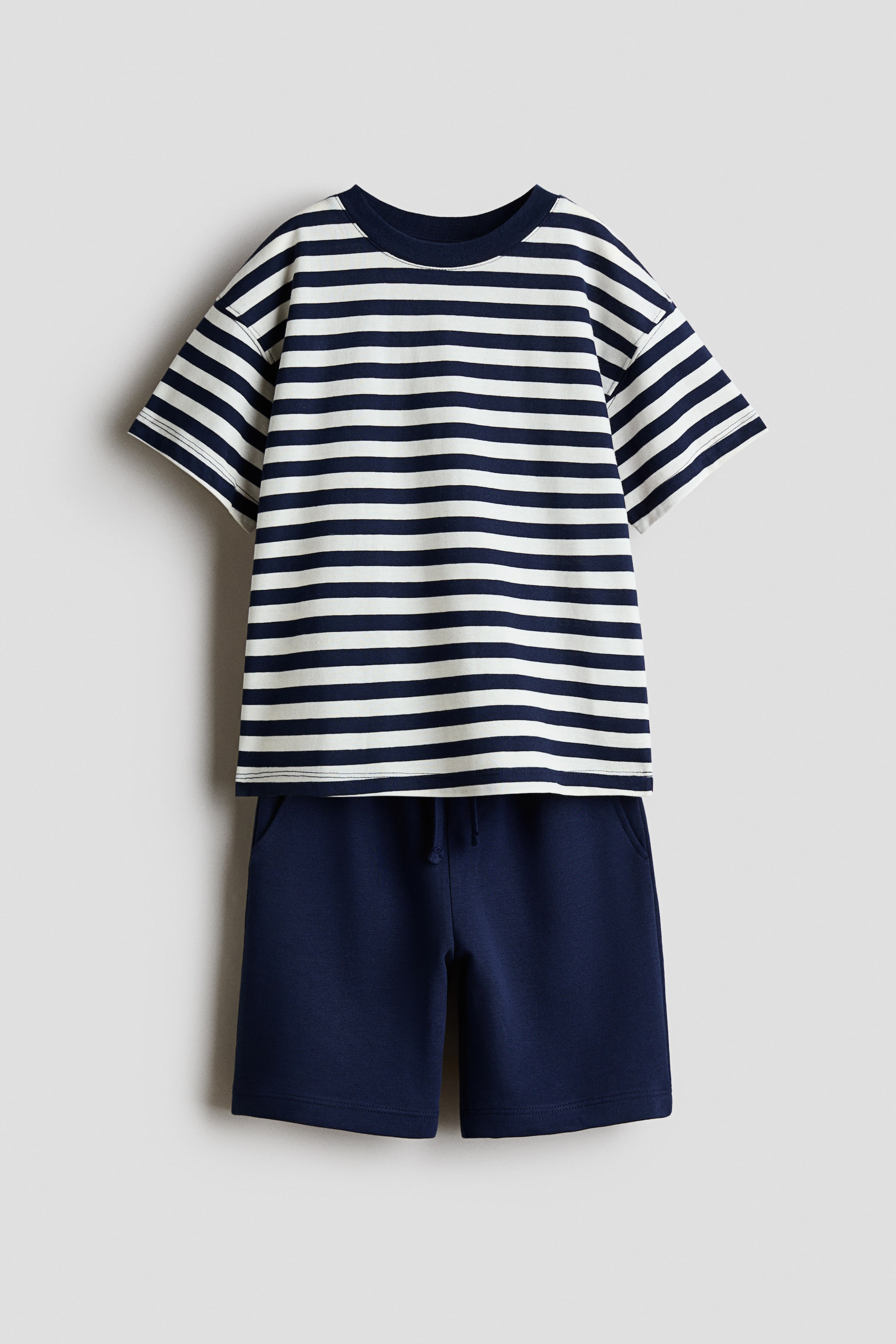 View larger image: A two-piece children's clothing set is shown from the front, featuring a short-sleeved T-shirt with navy blue and white horizontal stripes and a solid navy ribbed crew neck. The matching solid navy blue shorts feature an elastic waistband with drawstrings and side pockets.