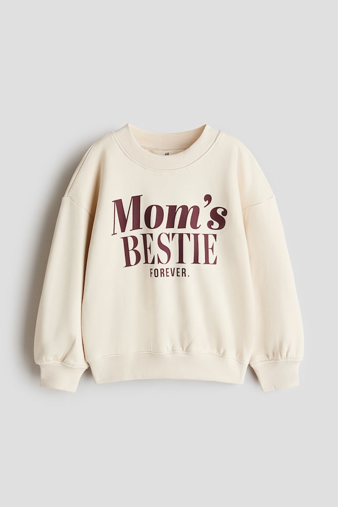 Ein cremefarbenes Sweatshirt in lockerer Passform ist von vorne zu sehen und prÃ¤sentiert einen runden, gerippt eingefassten Halsausschnitt, Ã¼berschnittene Schultern sowie gerippte BÃ¼ndchen und einen gerippten Saum. Auf der Brust ist ein burgunderfarbener Schriftzug mit der Aufschrift âMom's BESTIE FOREVERâ zu sehen. - 1