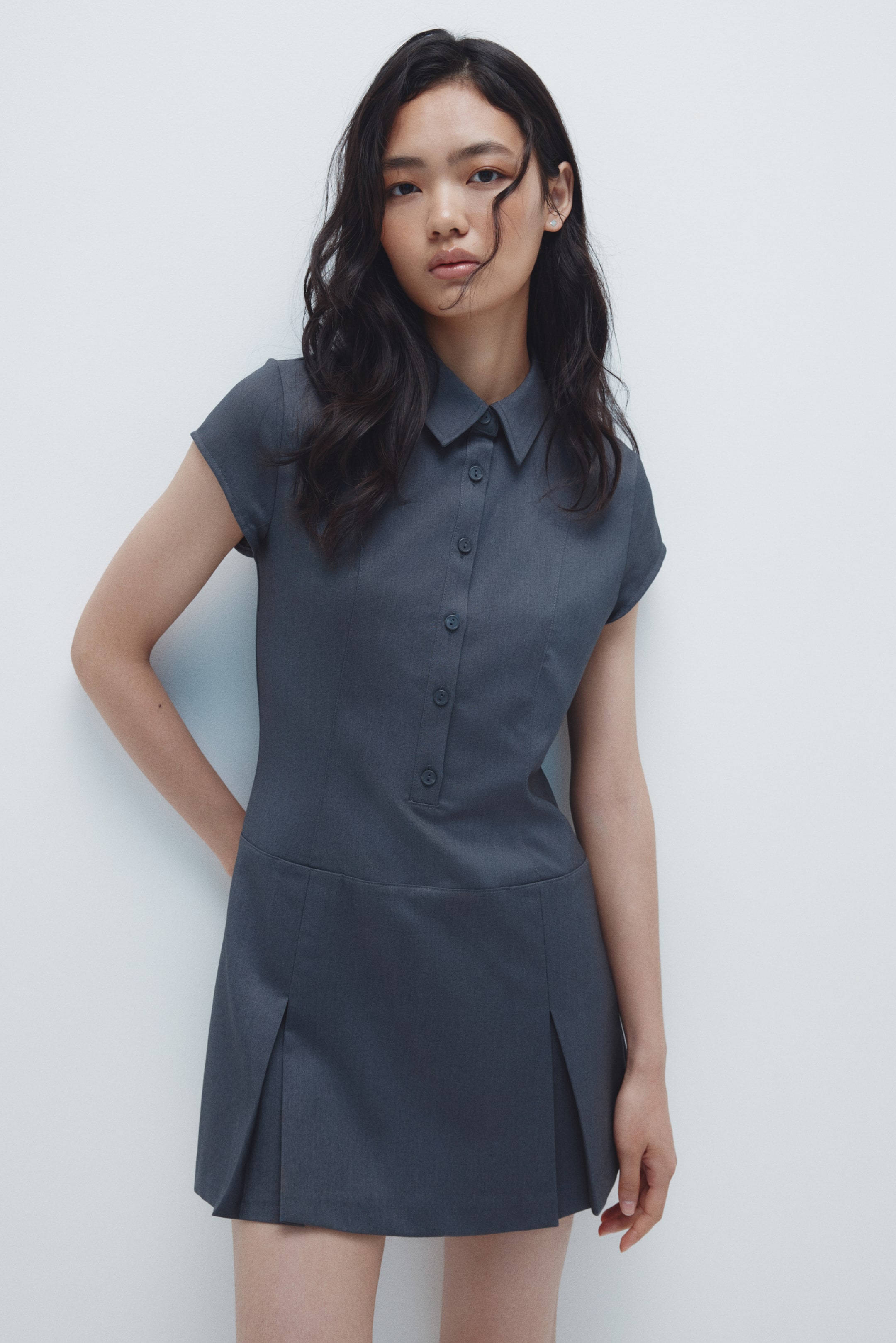 拡大画像を表示: 暗灰色のドレスがフロントから紹介されています。特徴は、尖った襟と短いキャップスリーブです。ドレスは前面にボタンで留められるプラケットがあり、くっきりしたウエストラインがプリーツの入ったスカートにつながっています。