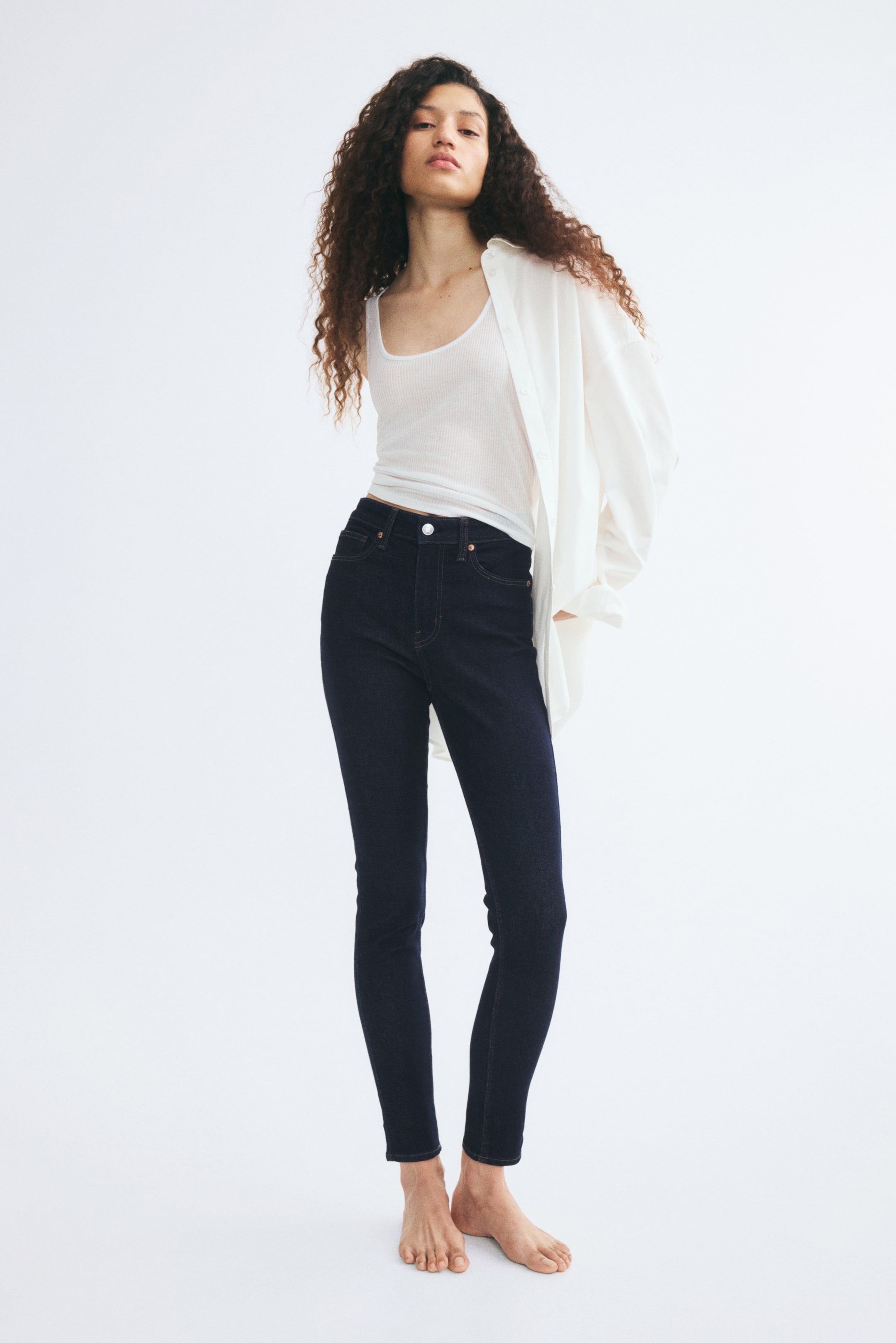 Büyük resmi görüntüle: Model, klasik beş cepli tasarıma ve bileklere doğru daralan slim fit kesime sahip koyu mavi, yüksek bel skinny jean giyiyor. Jean, beyaz ribana atlet ve açık bırakılmış, oversize beyaz düğmeli gömlek ile kombinlenmiş.