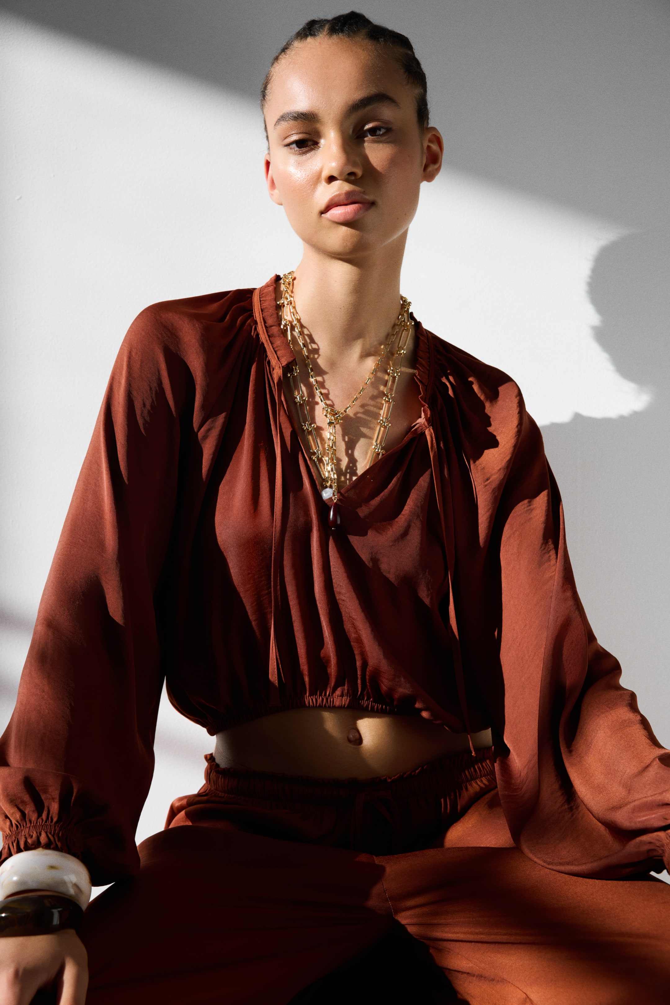 View larger image: A rust-colored blouse features a gathered neckline with ties, voluminous long sleeves gathered at the cuffs, and a cropped elasticated hem that reveals the midriff. Matching rust-colored pants are visible below the blouse, while layered gold-toned necklaces adorn the blouse's neckline, and two wide bracelets encircle one wrist.