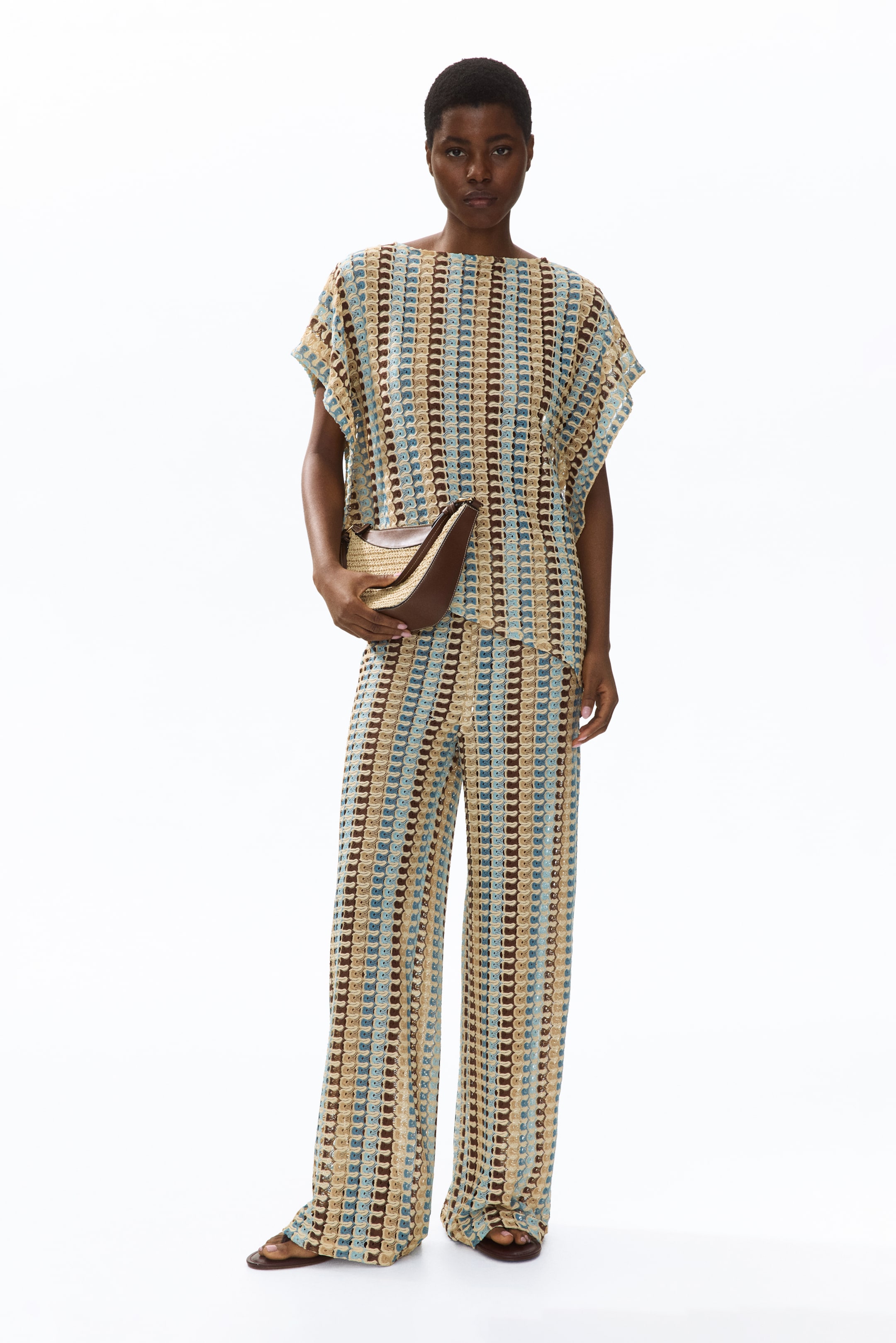 View larger image: A coordinated crochet-knit two-piece set includes a short-sleeve boat neck top and wide-leg pants. The set features a repeating geometric pattern in vertical stripes of brown, light blue, and beige, complemented by a woven straw and brown leather handbag.